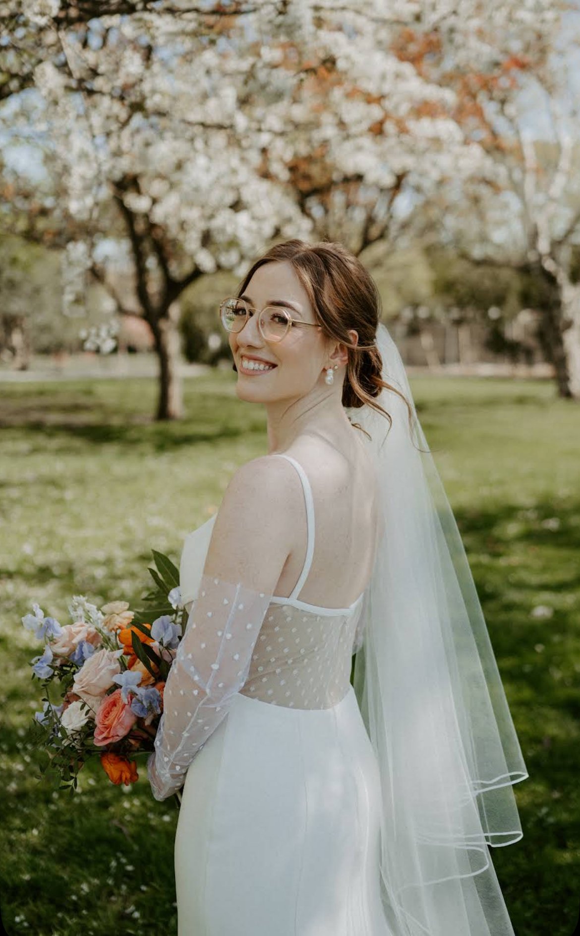 Bride in a white wedding dress with polka dot sleeves holding a colorful bouquet, standing outdoors near blooming trees, smiling and wearing glasses.