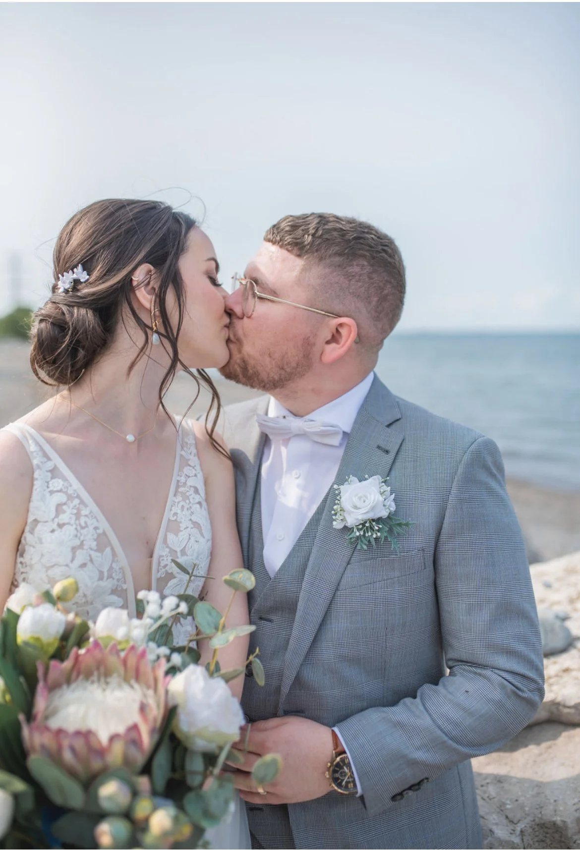 Bride and groom kissing at a beach with a bouquet, wearing a lace dress and gray suit.
