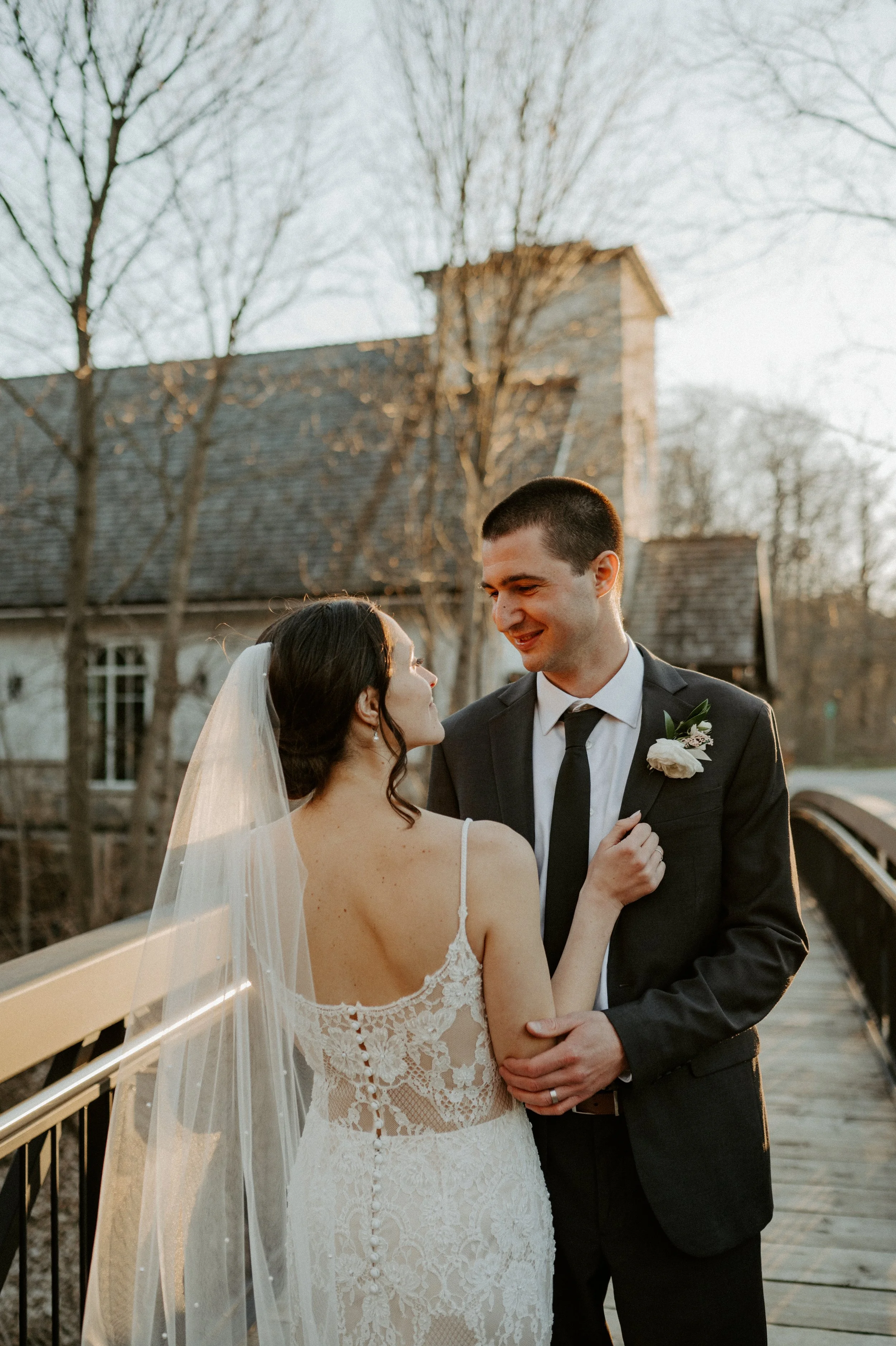 Bride and groom standing on a wooden bridge during their wedding, with the bride in a lace dress and veil, and the groom in a suit with a boutonniere.