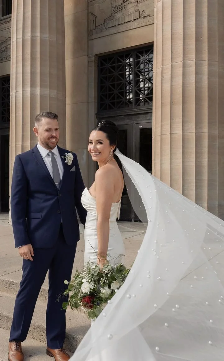 Bride and groom posing outside a building with large columns, bride holding a bouquet, wearing a long veil, groom in a navy suit.