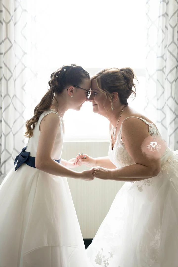 A woman in a wedding dress and a young girl in a white dress holding hands and touching foreheads in front of a bright window.