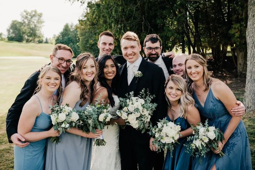 Group of people at a wedding, including a bride and groom, surrounded by bridesmaids in blue dresses and groomsmen in suits, holding floral bouquets, standing outdoors.