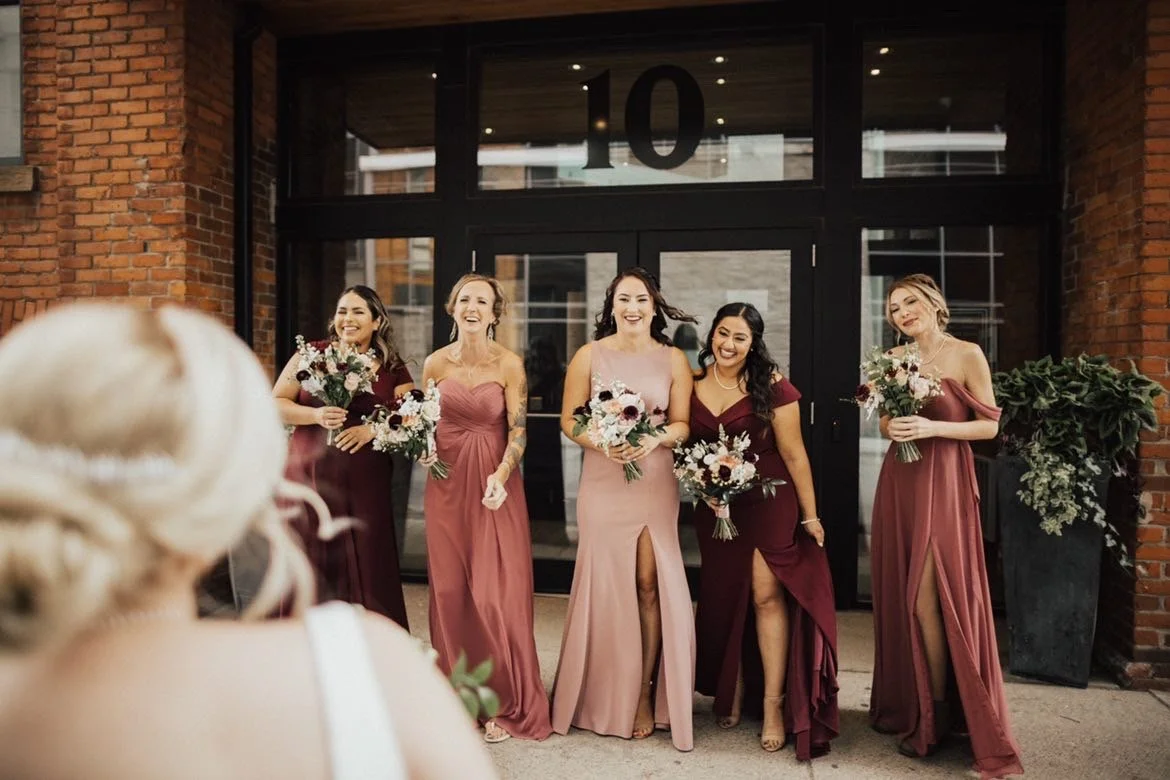 Group of bridesmaids in maroon and blush dresses holding bouquets, smiling in front of brick building entrance with number 10.