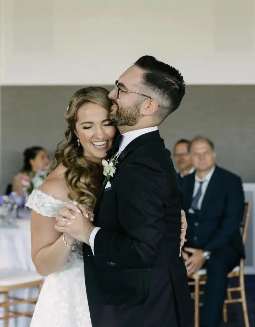 Bride and groom smiling while dancing at their wedding reception, with guests in the background.