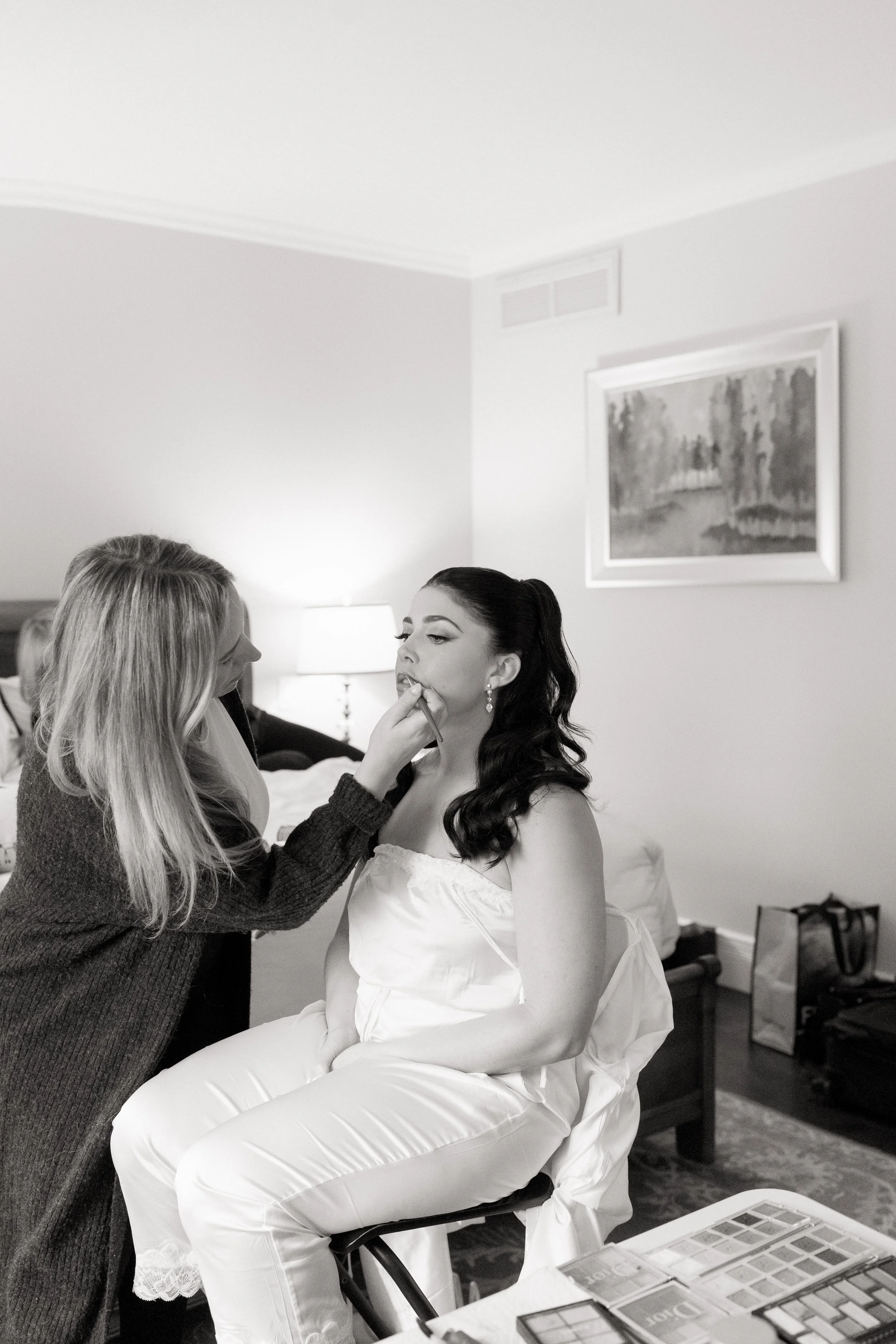 A woman getting her makeup done while seated on a chair, with another woman applying lipstick or lip gloss. The setting is a bedroom with a bed, nightstand with a lamp, a painting on the wall, and makeup products on a table.