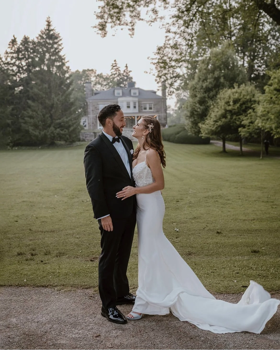 Bride and groom in formal attire standing in a garden with trees and a large house in the background.