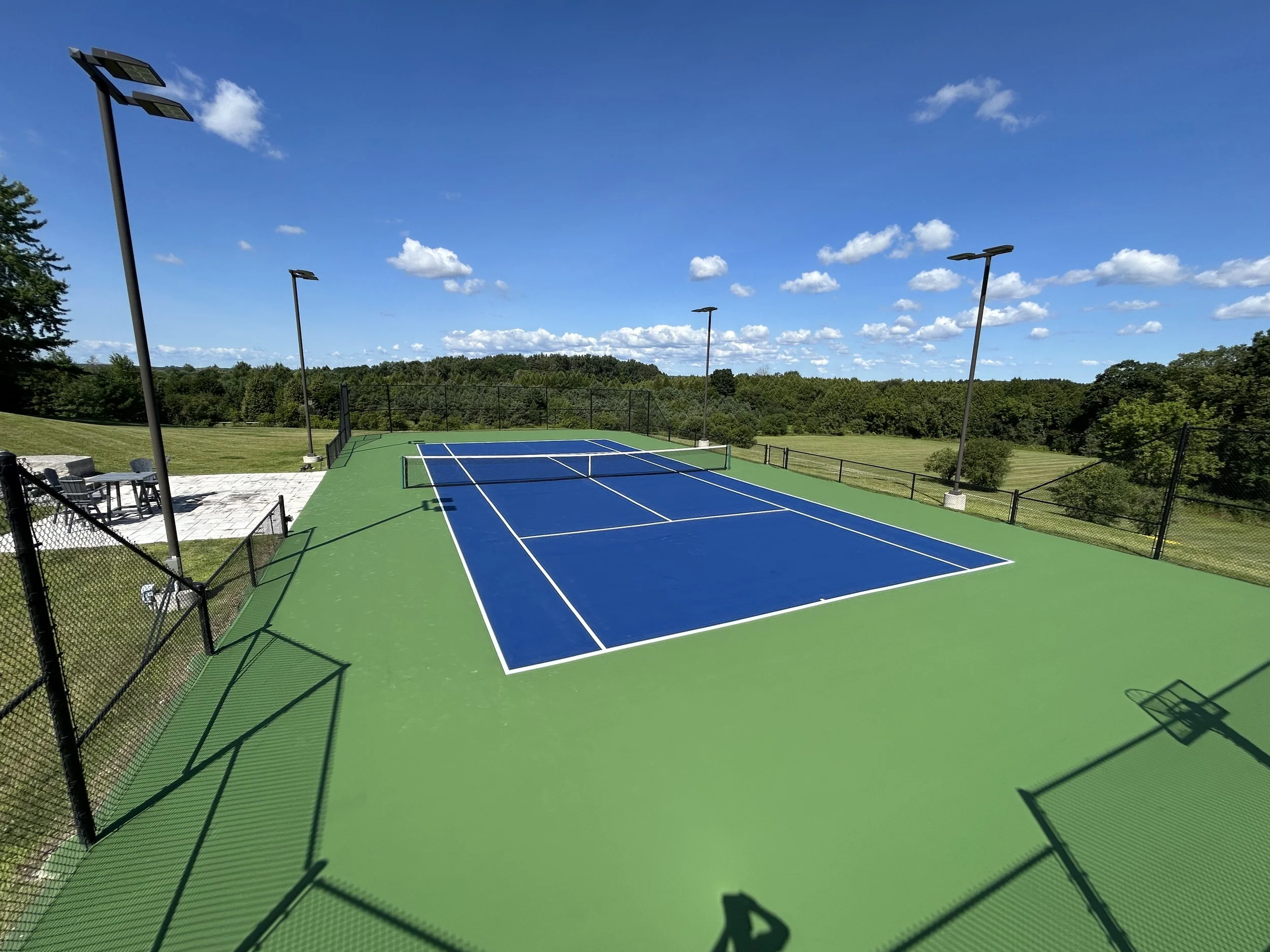 A tennis court with blue surface and green surroundings, surrounded by a black fence, under a bright blue sky with scattered white clouds, and tall light poles for nighttime play.