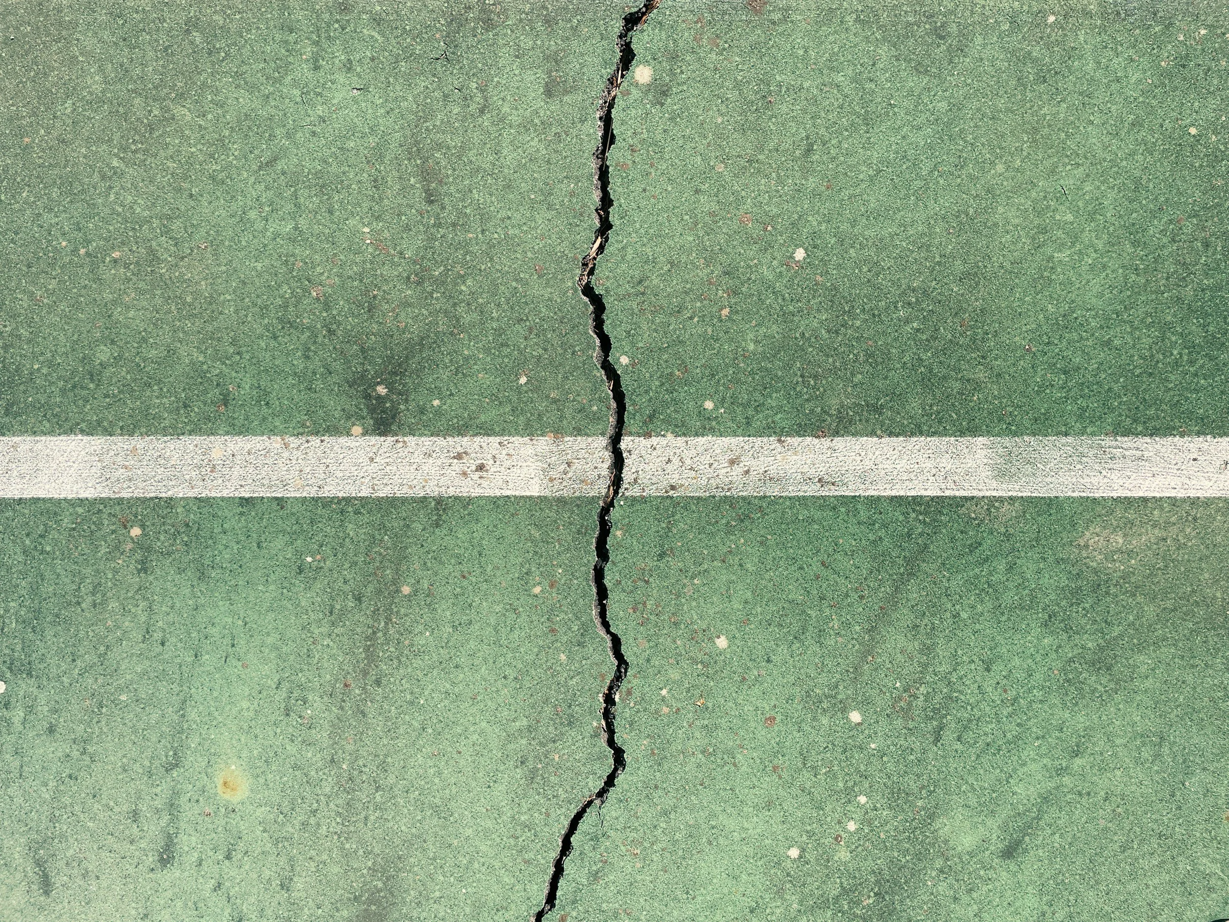 A close-up of a green sports court surface with a large crack running through the middle, and a white line at the bottom edge.
