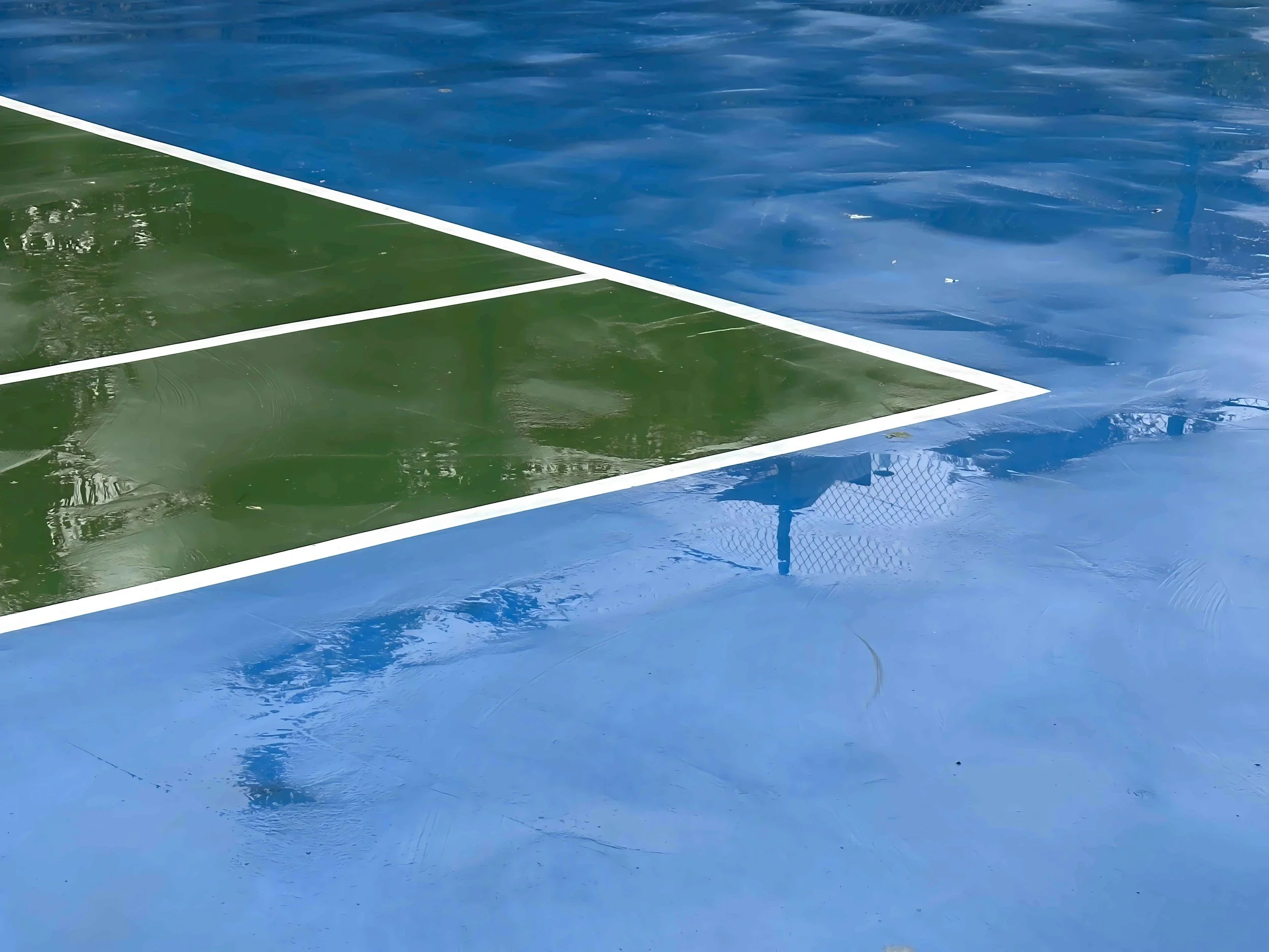 A tennis court with a green and blue surface, water pooling on parts of it, and visible white boundary lines.