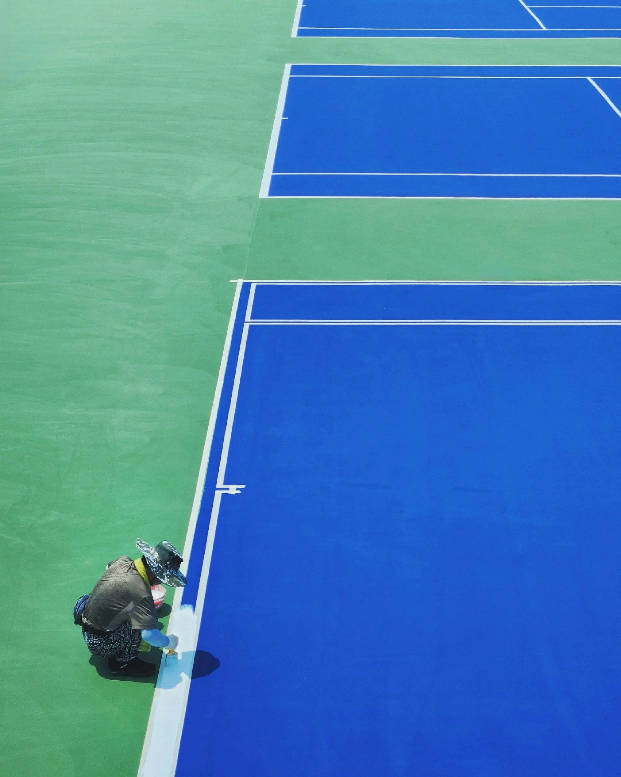 A person wearing a baseball cap, hoodie, and painting a blue line on a green tennis court, with paint supplies nearby.