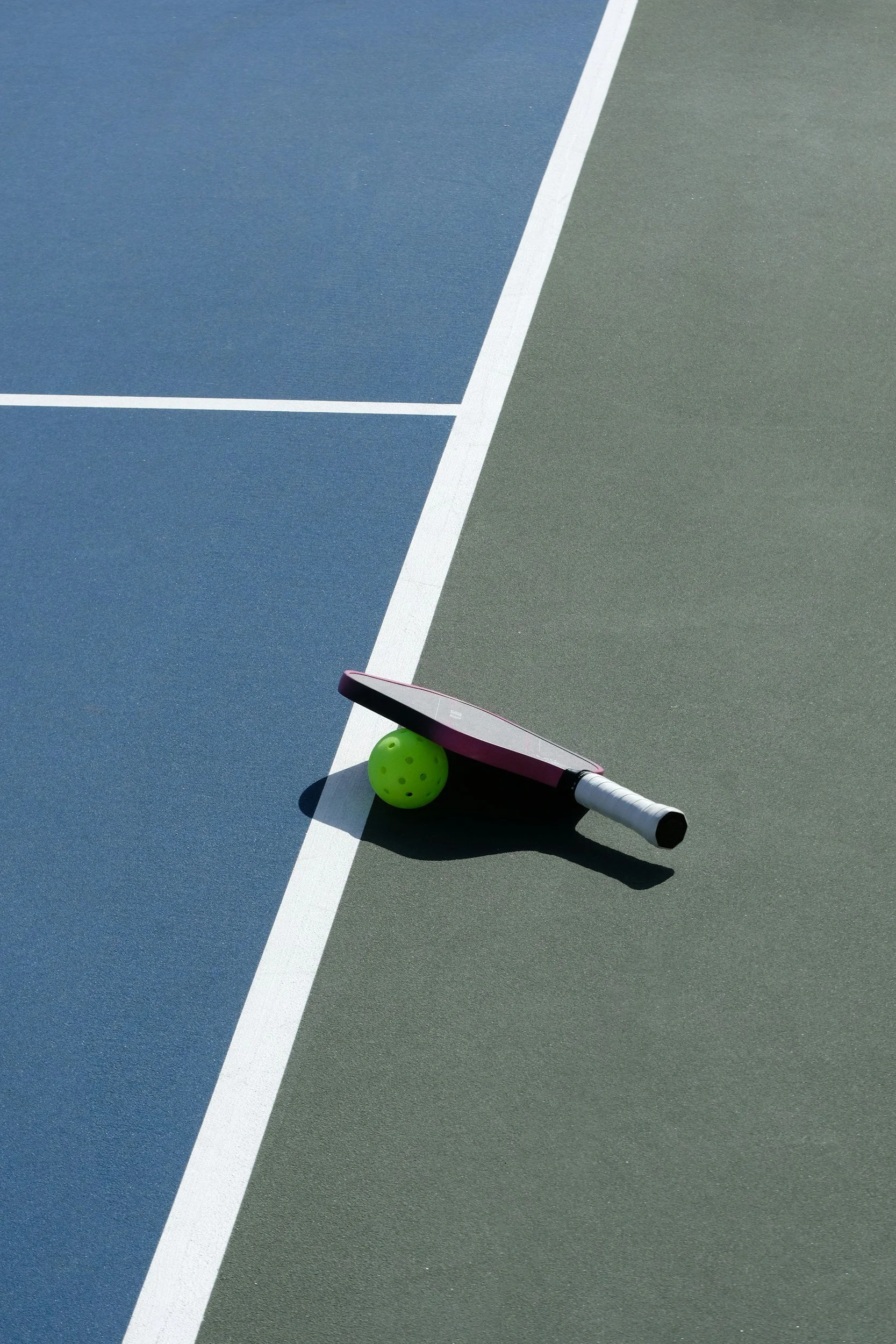 Part of a tennis court with a tennis racket, green tennis ball, and a small pink container on the ground.