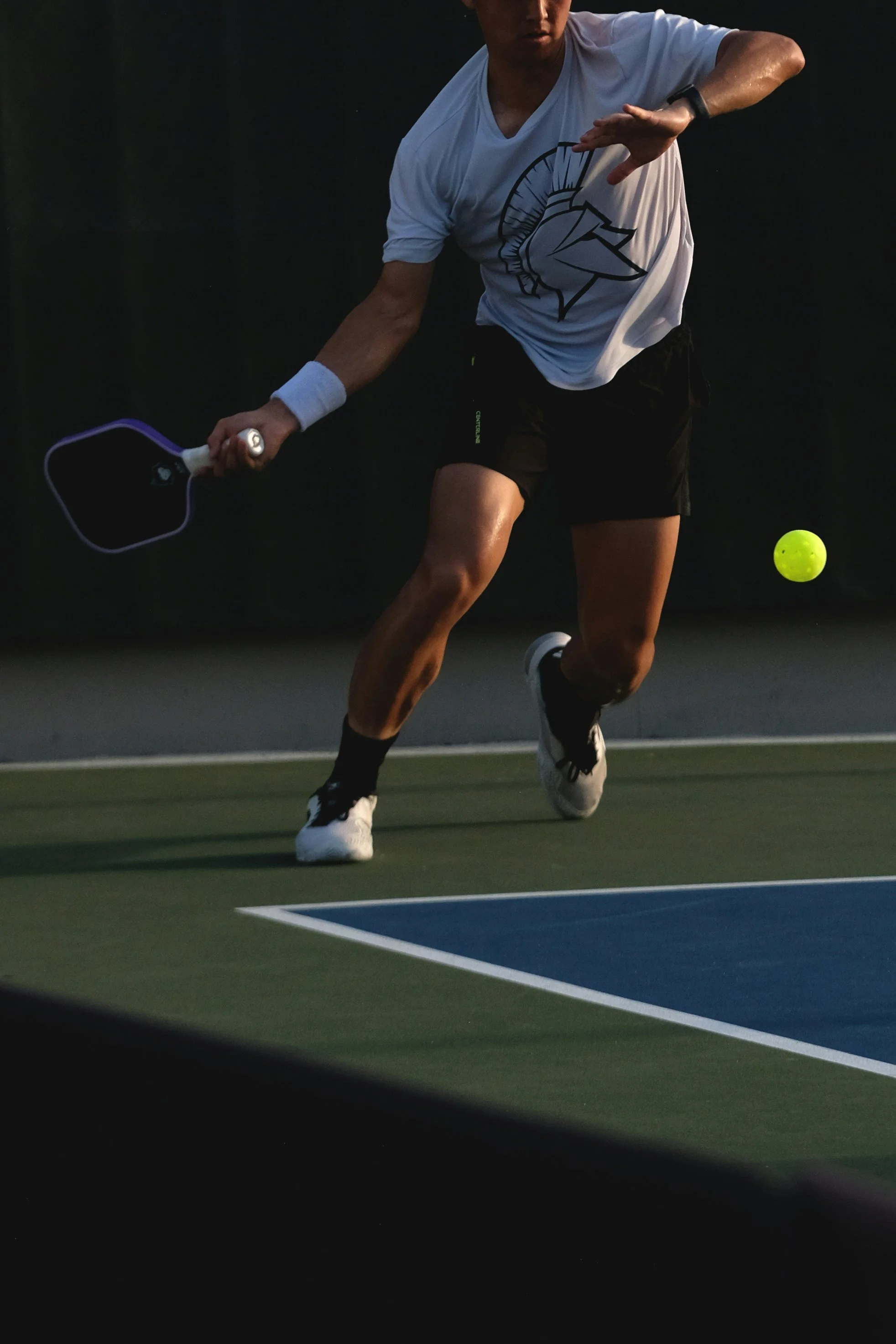 A man playing tennis on an indoor court, wearing a white T-shirt, black shorts, and tennis shoes, with a yellow tennis ball in motion near his right leg.
