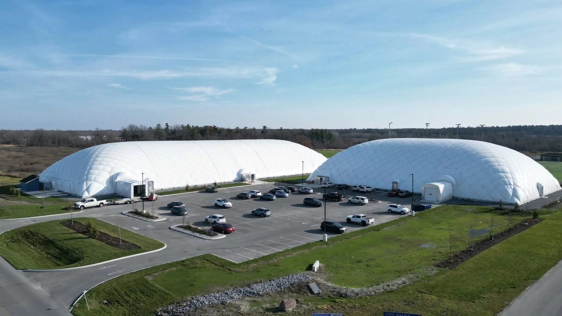 Two large white dome structures with car parking lot in front, surrounded by green grass and trees under a blue sky.