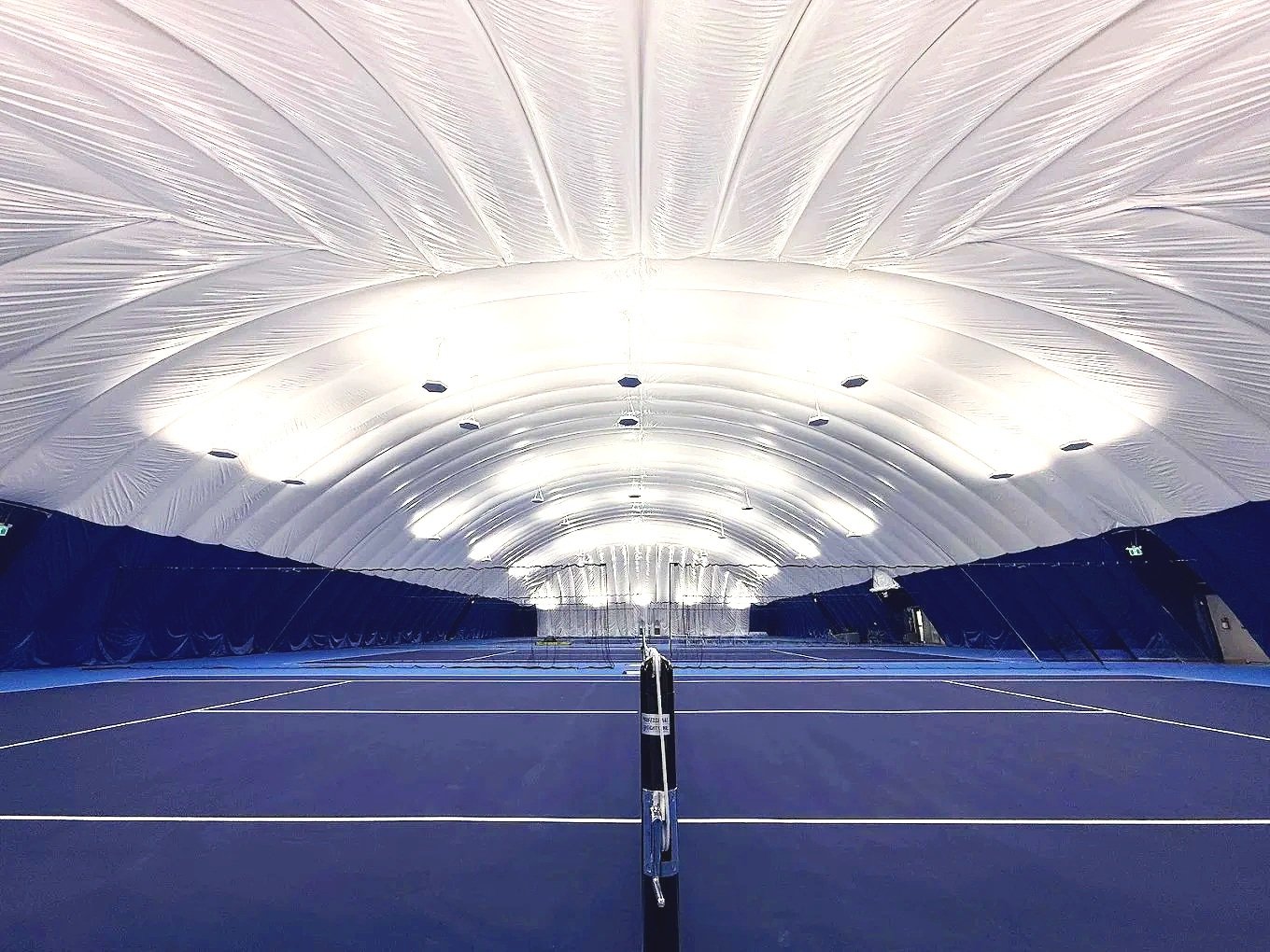 An indoor tennis court with a blue playing surface, illuminated by bright white light fixtures, under a white, curved, fabric ceiling.