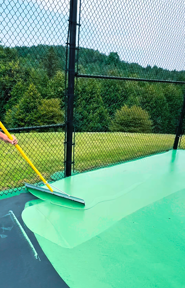Person applying green pool paint or coating onto a surface with a roller, outdoor setting with a chain-link fence and trees in the background.