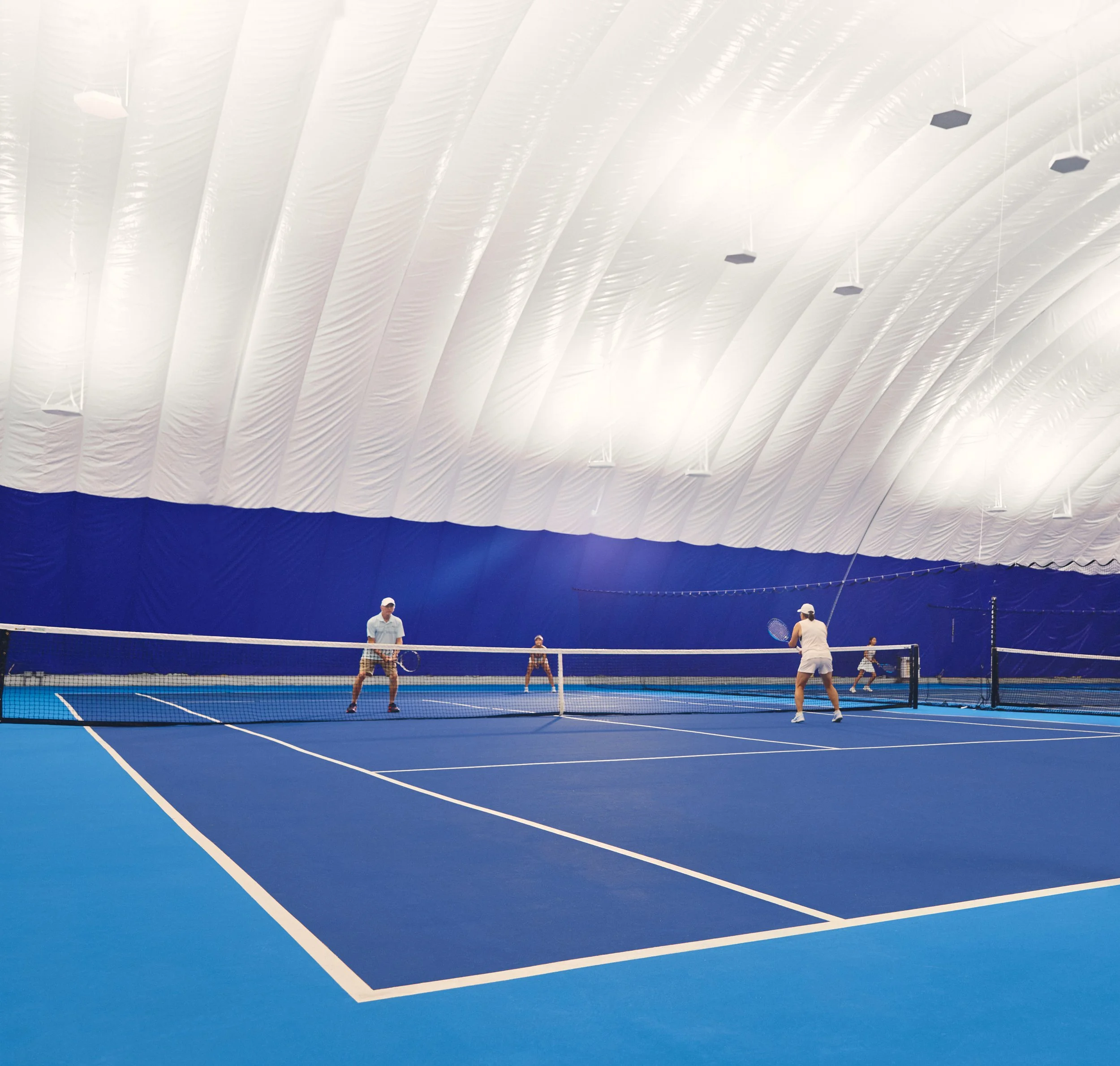 Indoor tennis court with players, a high ceiling with long white panels, and hanging lights.