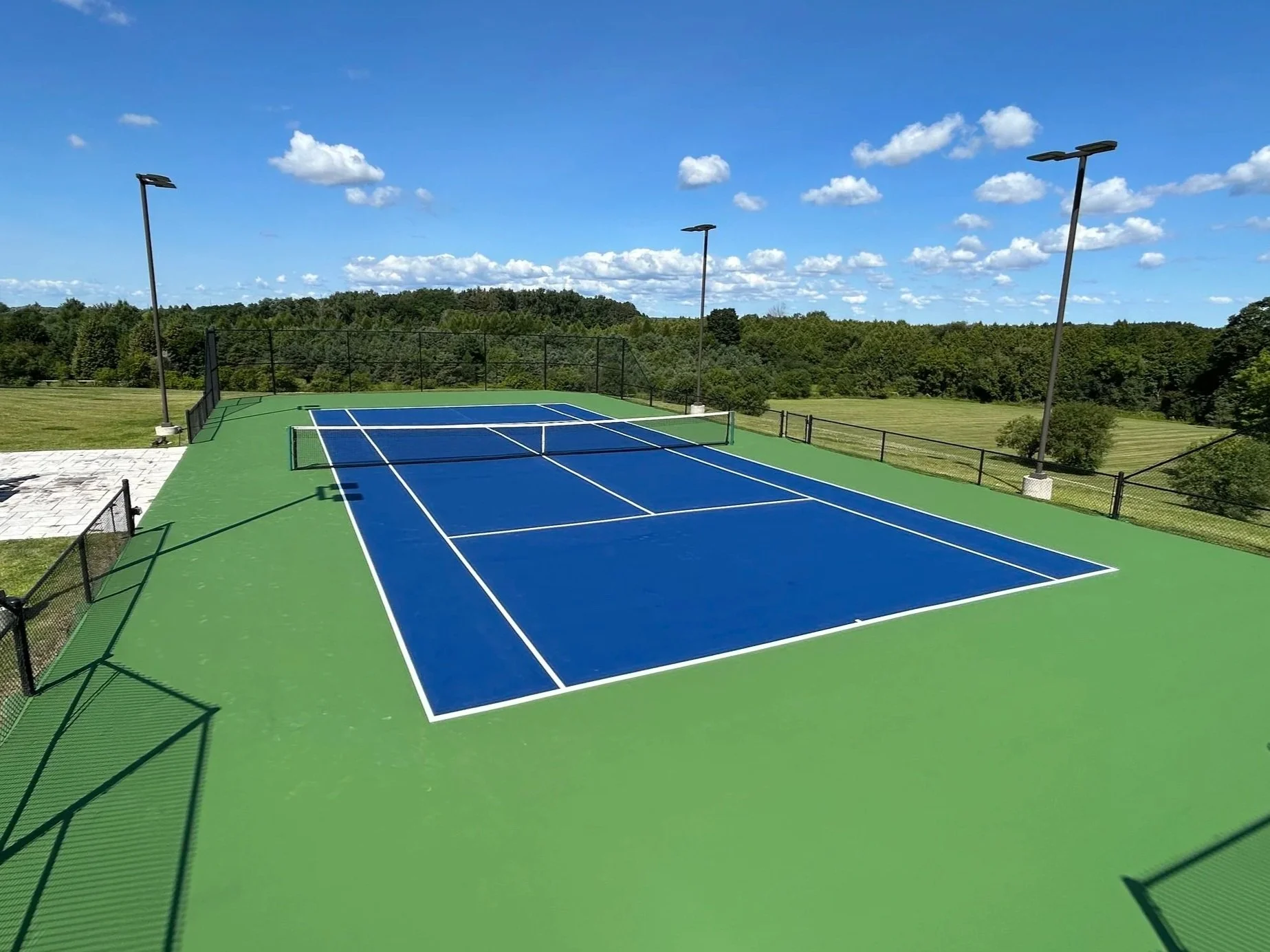 An empty outdoor tennis court with a blue surface and white lines, surrounded by a black fence, under a clear blue sky with scattered clouds, and green trees in the background.