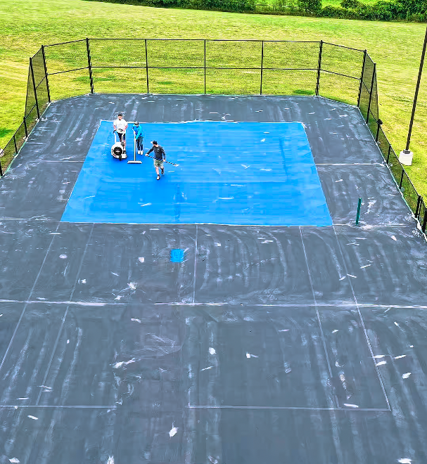 Two workers cleaning a tennis court with a power washer, surrounded by a black fence, with green lawn and trees in the background.