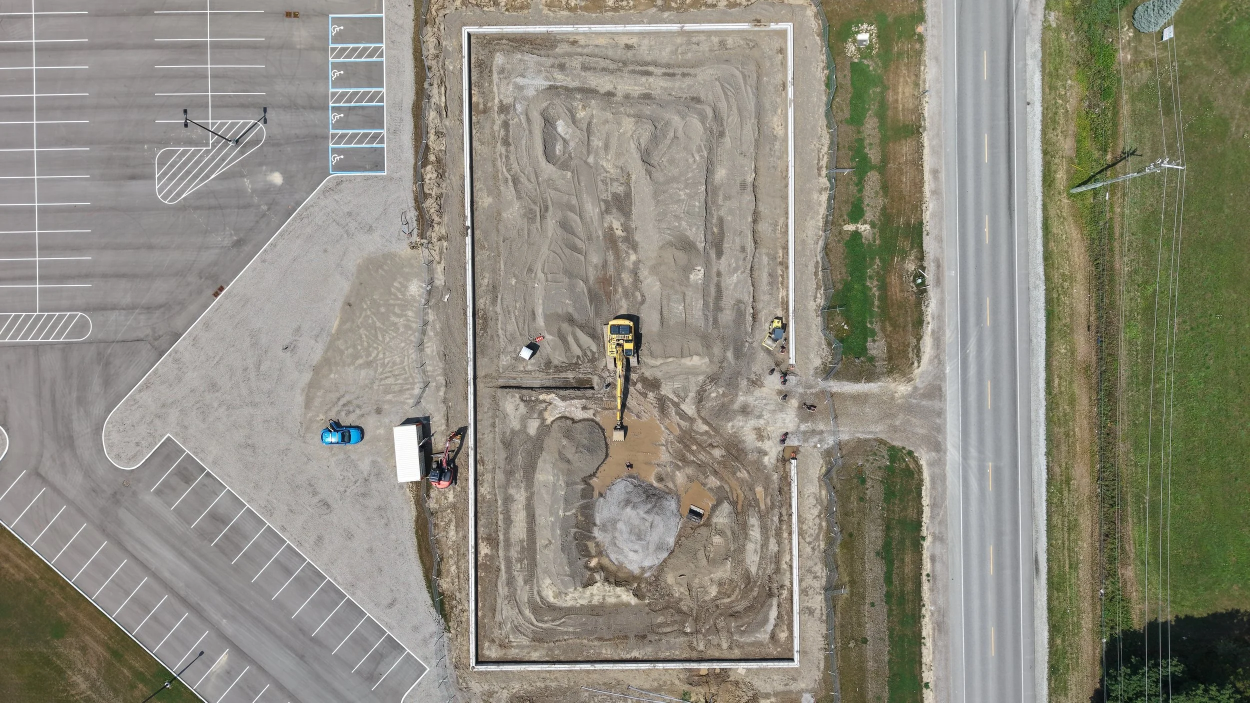 An aerial view of a construction site with an excavator digging, surrounded by a newly poured concrete foundation. To the left, there is a parking lot with marked parking spaces and handicap spaces. On the right, a paved road runs parallel to the construction area, with utility poles and wires alongside it.