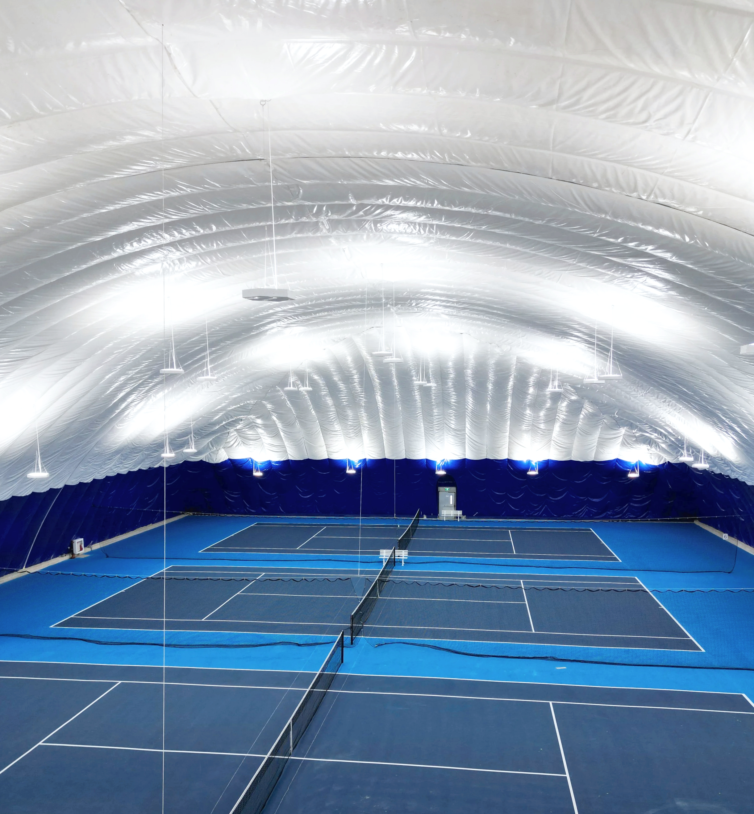 Indoor tennis court with blue surface, white boundary lines, black net, and blue curtains. The ceiling is curved, white, and illuminated.