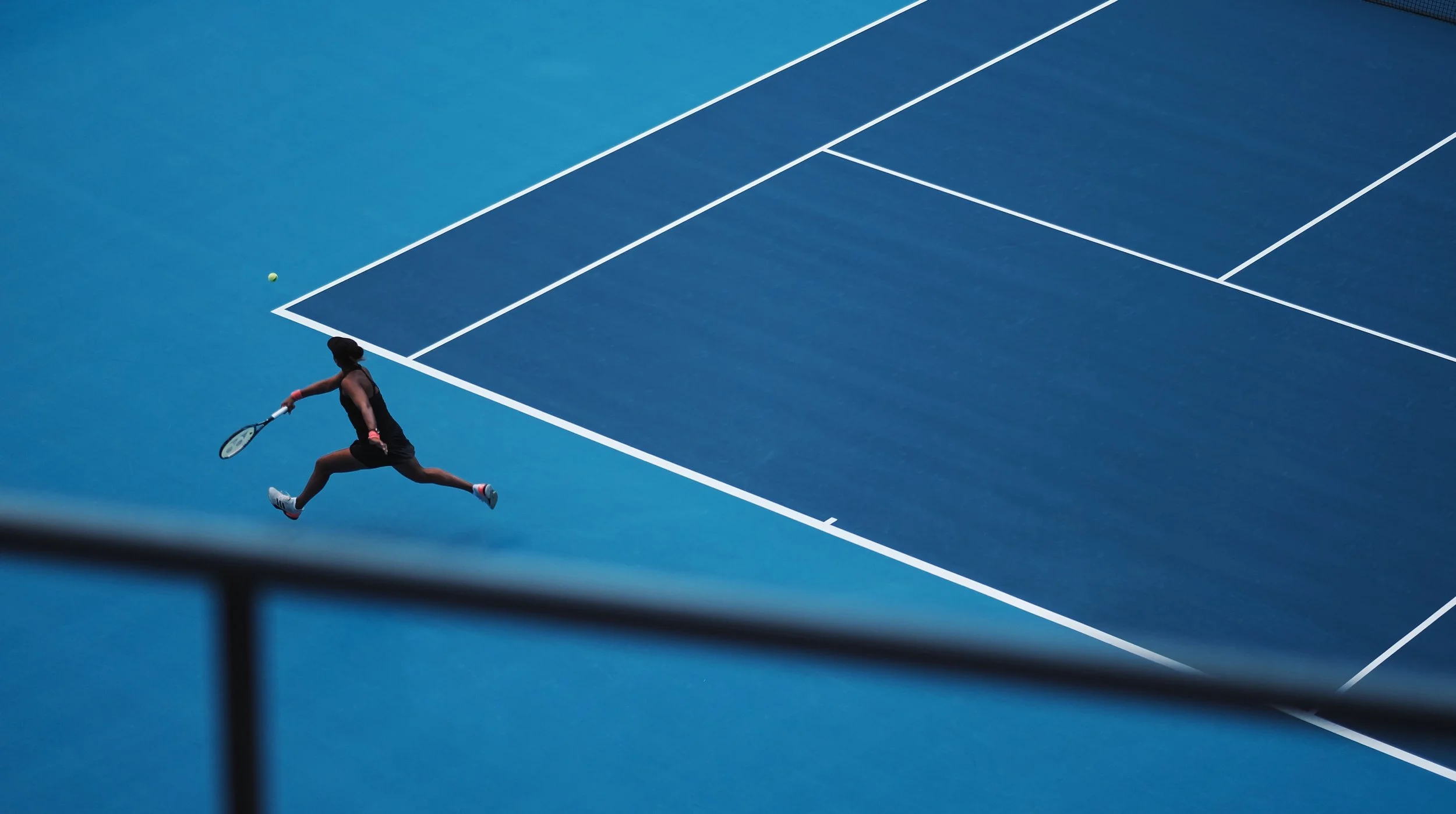 A female tennis player in black attire and white sneakers is jumping to hit a tennis ball on a blue tennis court with white lines.