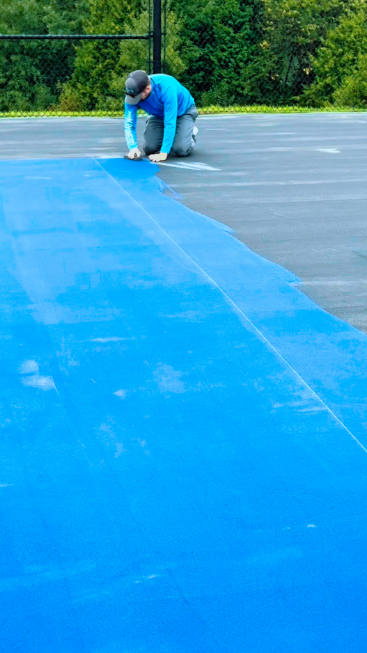 A person kneeling on an outdoor tennis court, painting or repairing a blue line on the court surface.