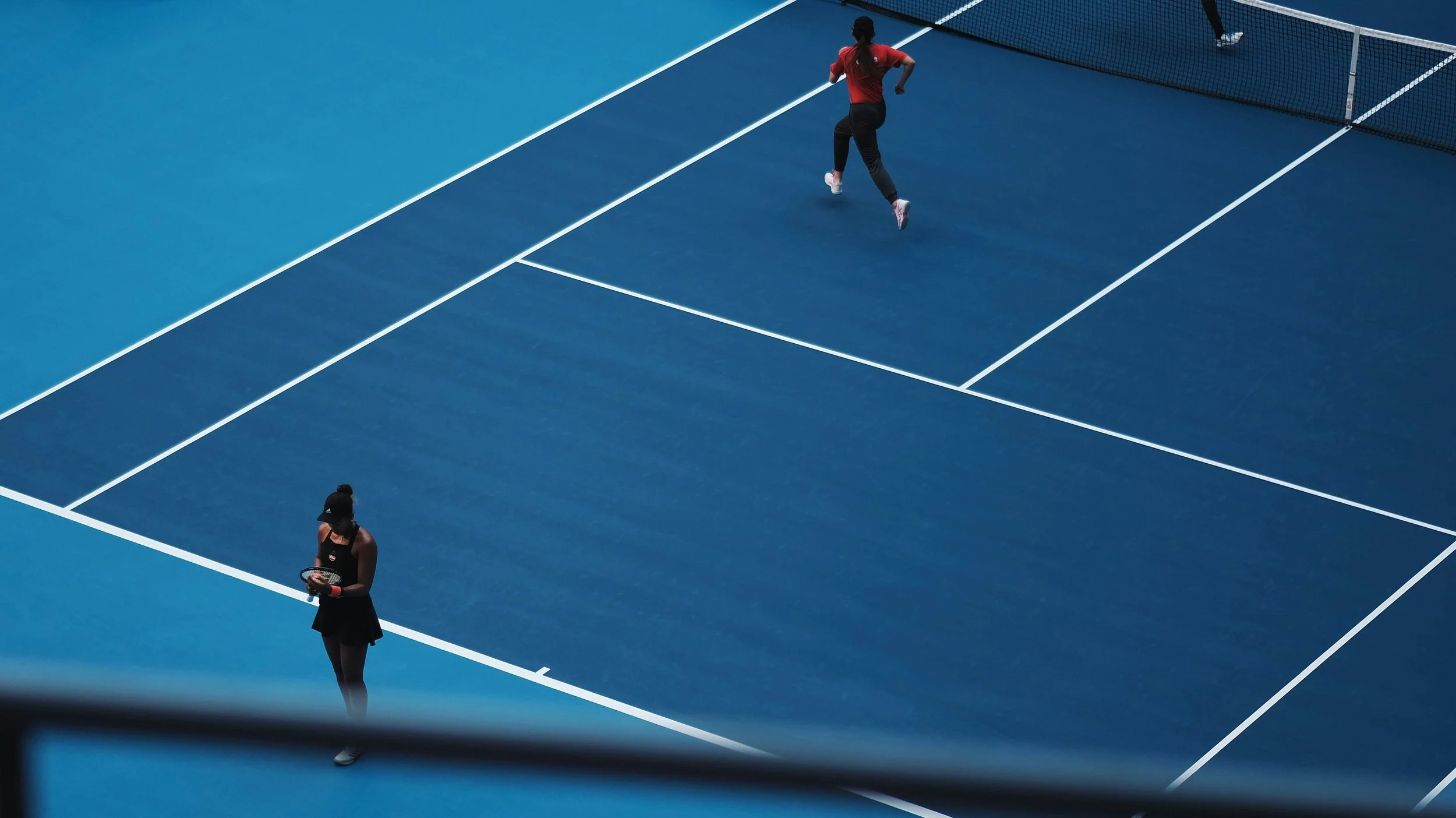 Two female tennis players on a blue tennis court, one near the net and the other farther back, both holding rackets, with a bow-shaped net in the background.