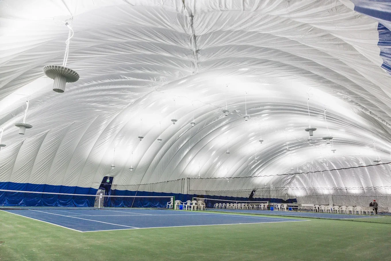 Indoor tennis court with a curved white fabric roof, blue walls, and chairs along the side.