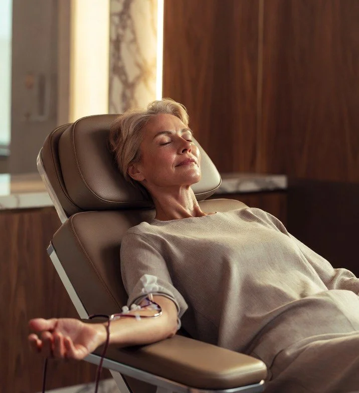 An older woman with short blonde hair donating blood in a medical facility, lying on a reclining chair with her eyes closed.