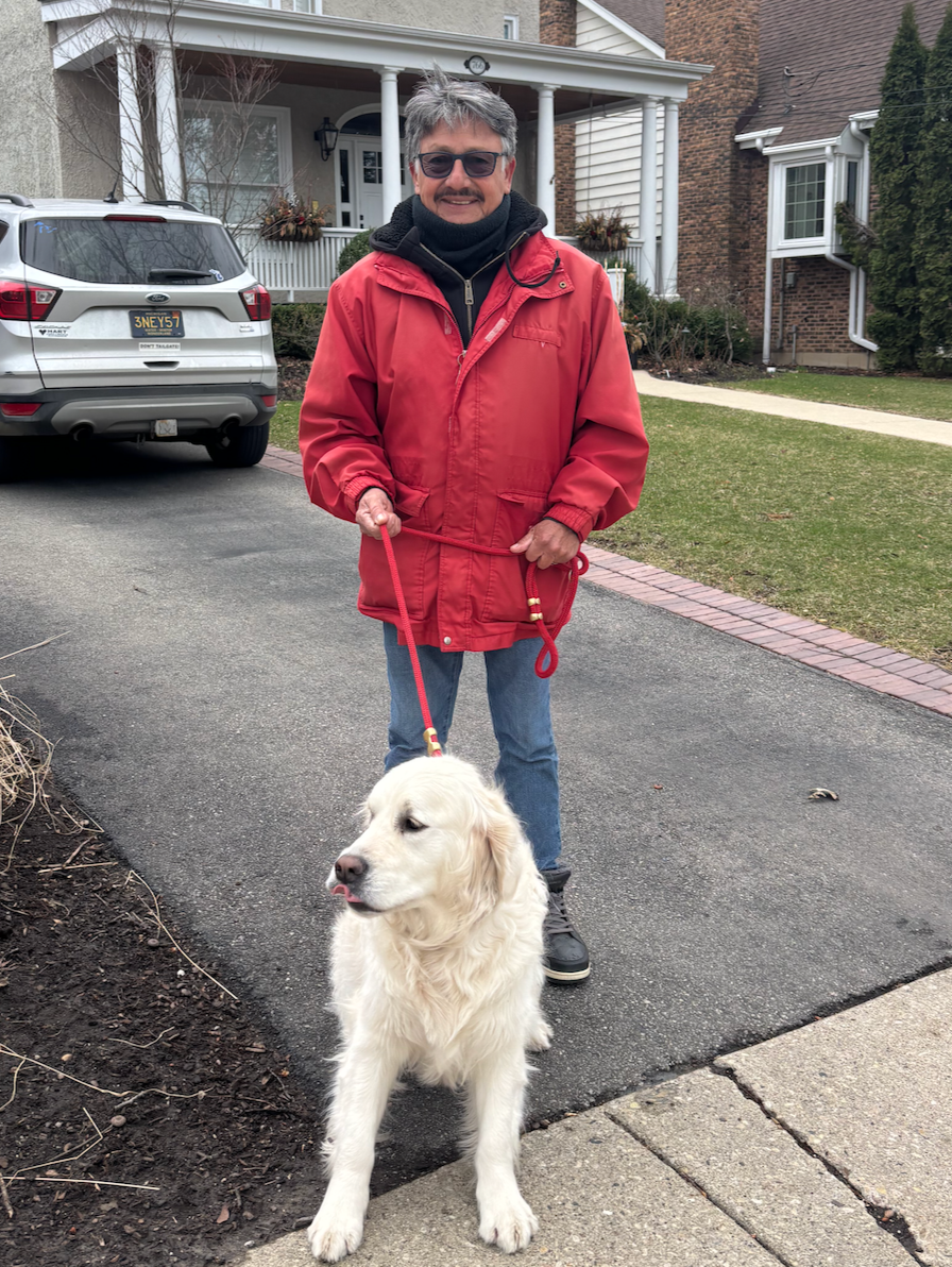 A dog walker in a red jacket stands in a residential driveway holding the leash of a cream-colored retriever, with homes and parked cars in the background.
