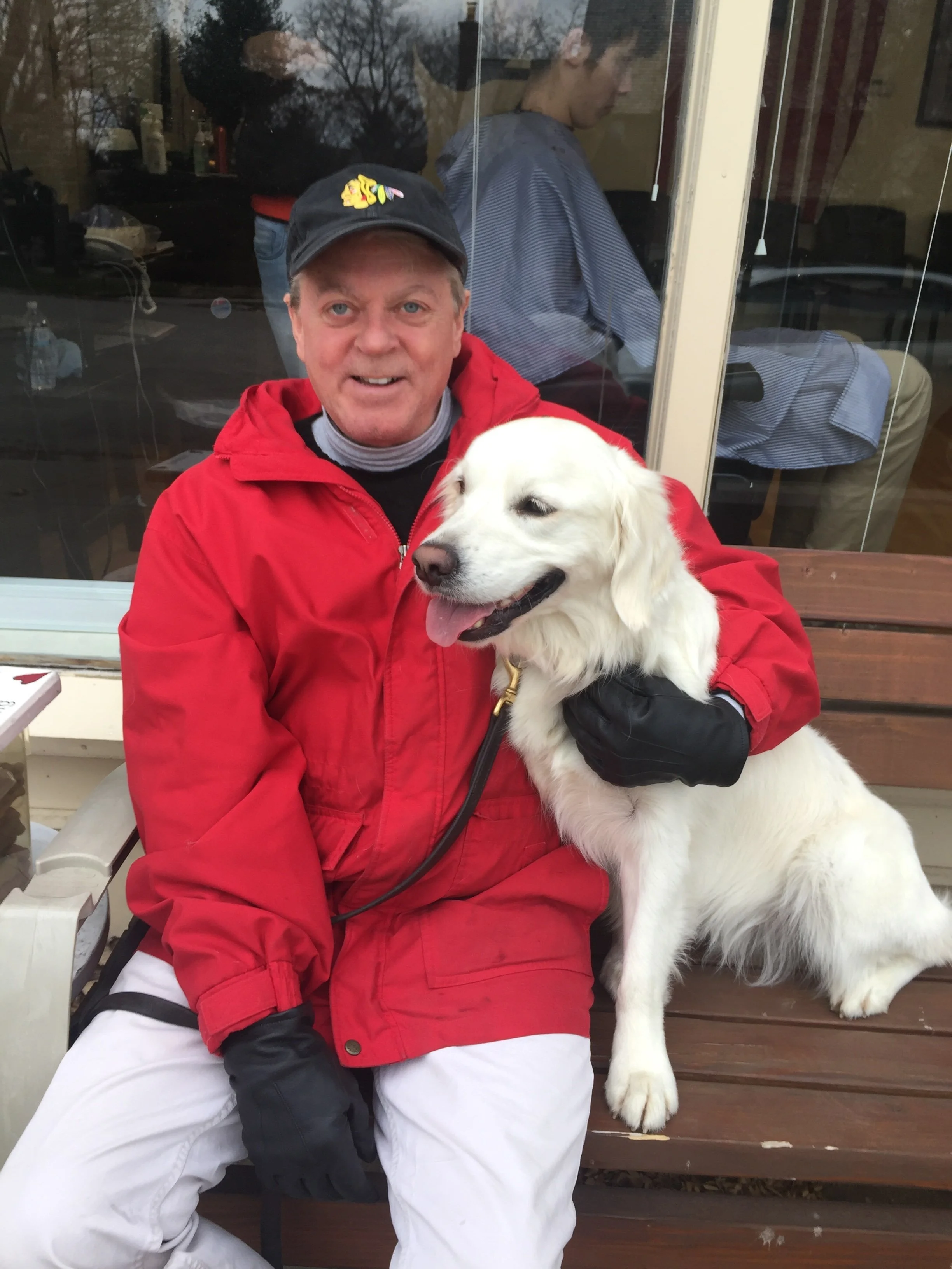 A man in a red jacket and black gloves sits on a bench with a happy white retriever at his side, outside a storefront window.