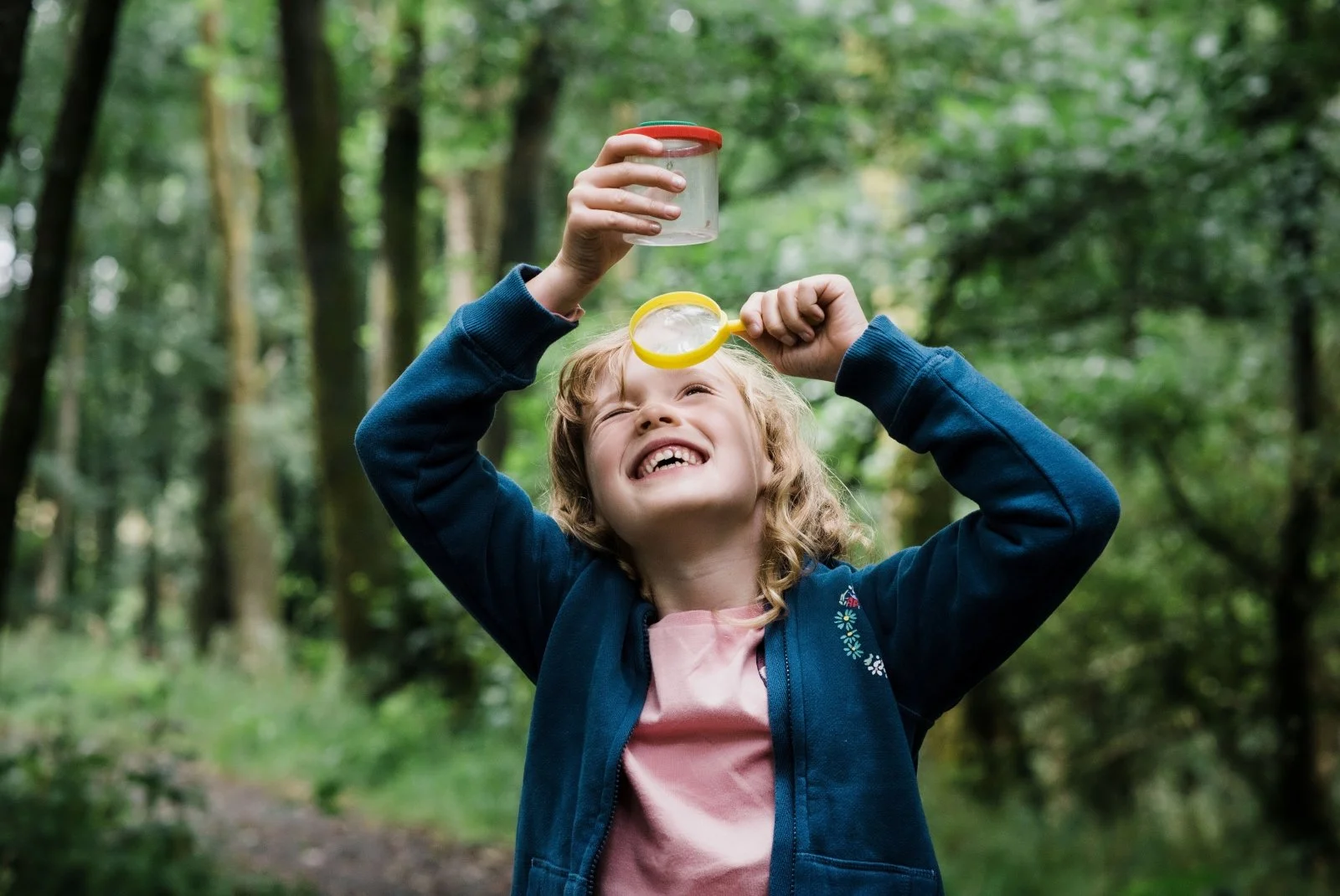 A young girl with curly blonde hair smiling and blowing bubbles outdoors in a forest.