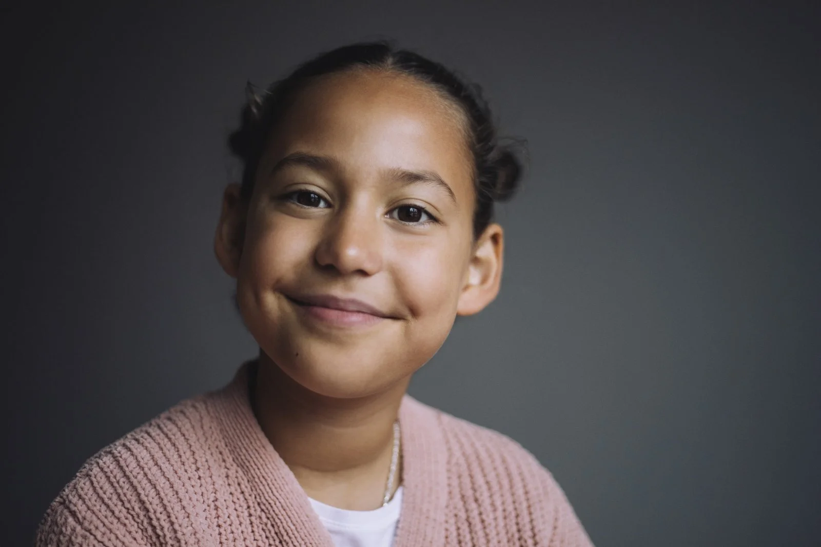 Portrait of a young girl with a warm smile, wearing a pink sweater and a white shirt, against a dark grey background.