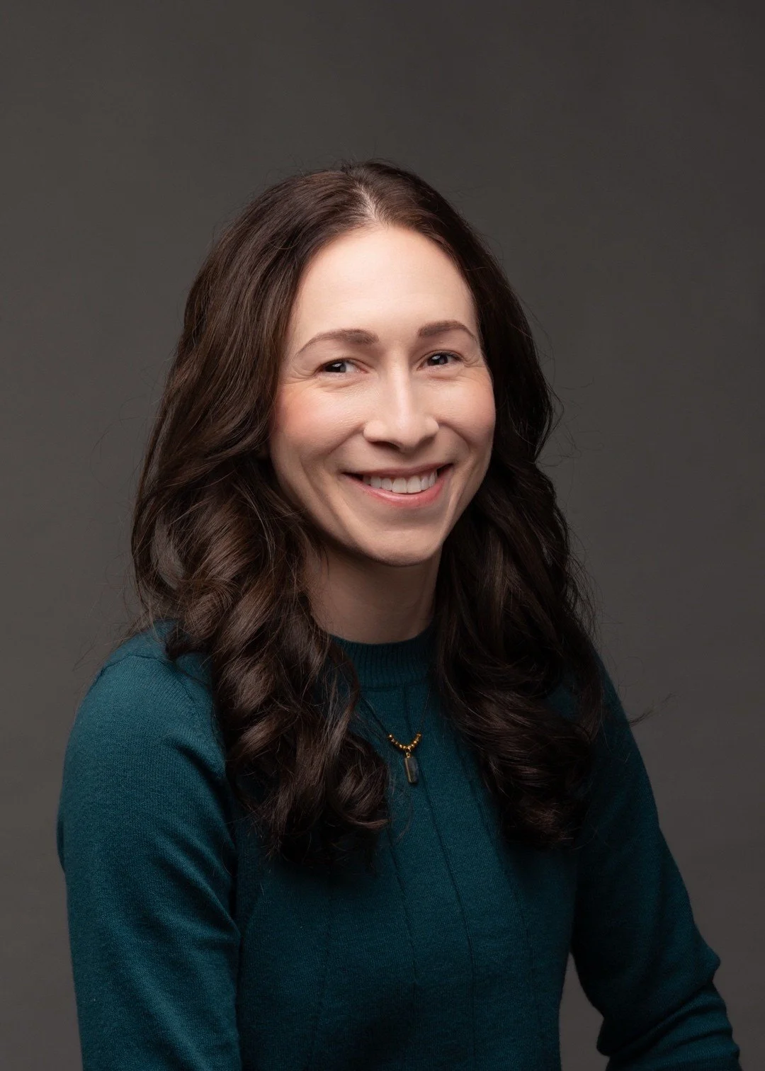 A woman with long brown hair smiling, wearing a teal sweater and a necklace with a pendant, against a gray background.