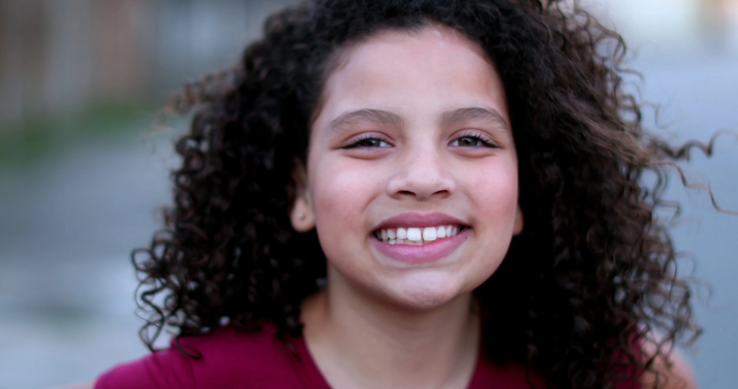 Close-up of a young girl with curly hair smiling outdoors.