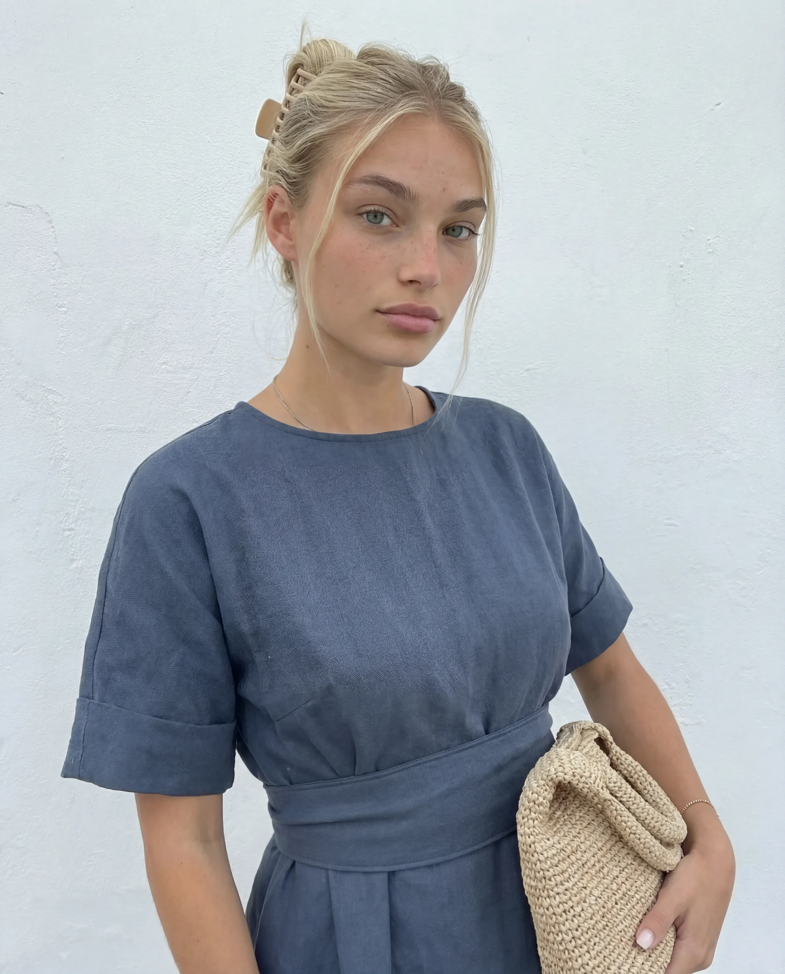 A young woman with blonde hair tied back, wearing a blue dress, holding a beige woven clutch, standing against a plain white wall.