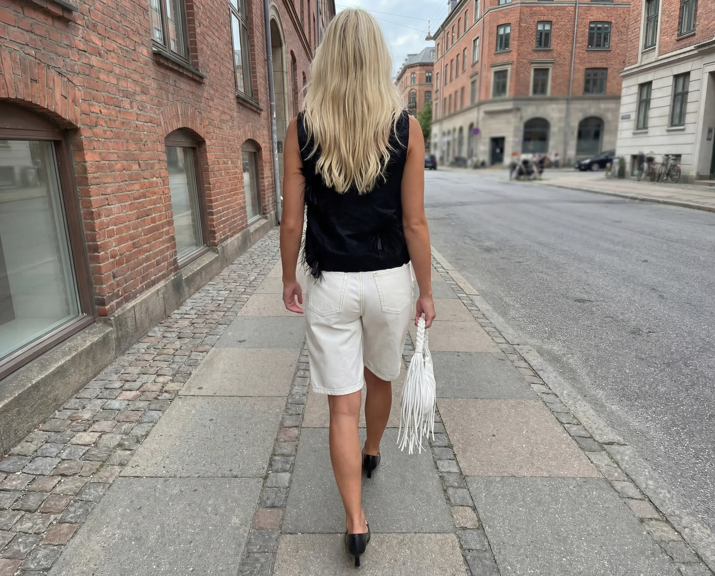 A woman walking down a city street with red brick buildings on either side, wearing a black sleeveless top, white shorts, and black heeled shoes, carrying a white fringed bag.