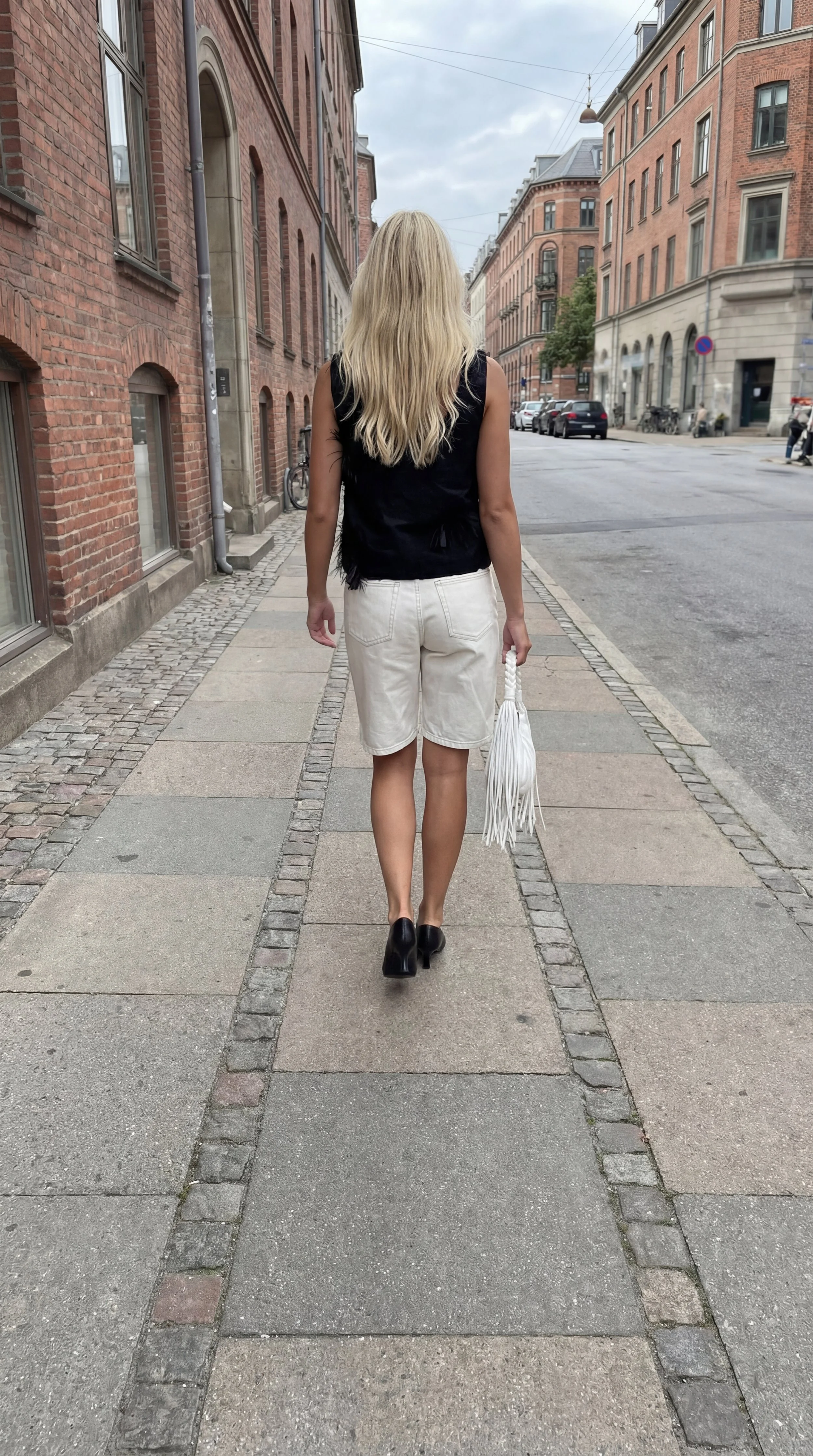 A woman walking down a city street with red brick buildings on either side, wearing a black sleeveless top, white shorts, and black heeled shoes, carrying a white fringed bag.