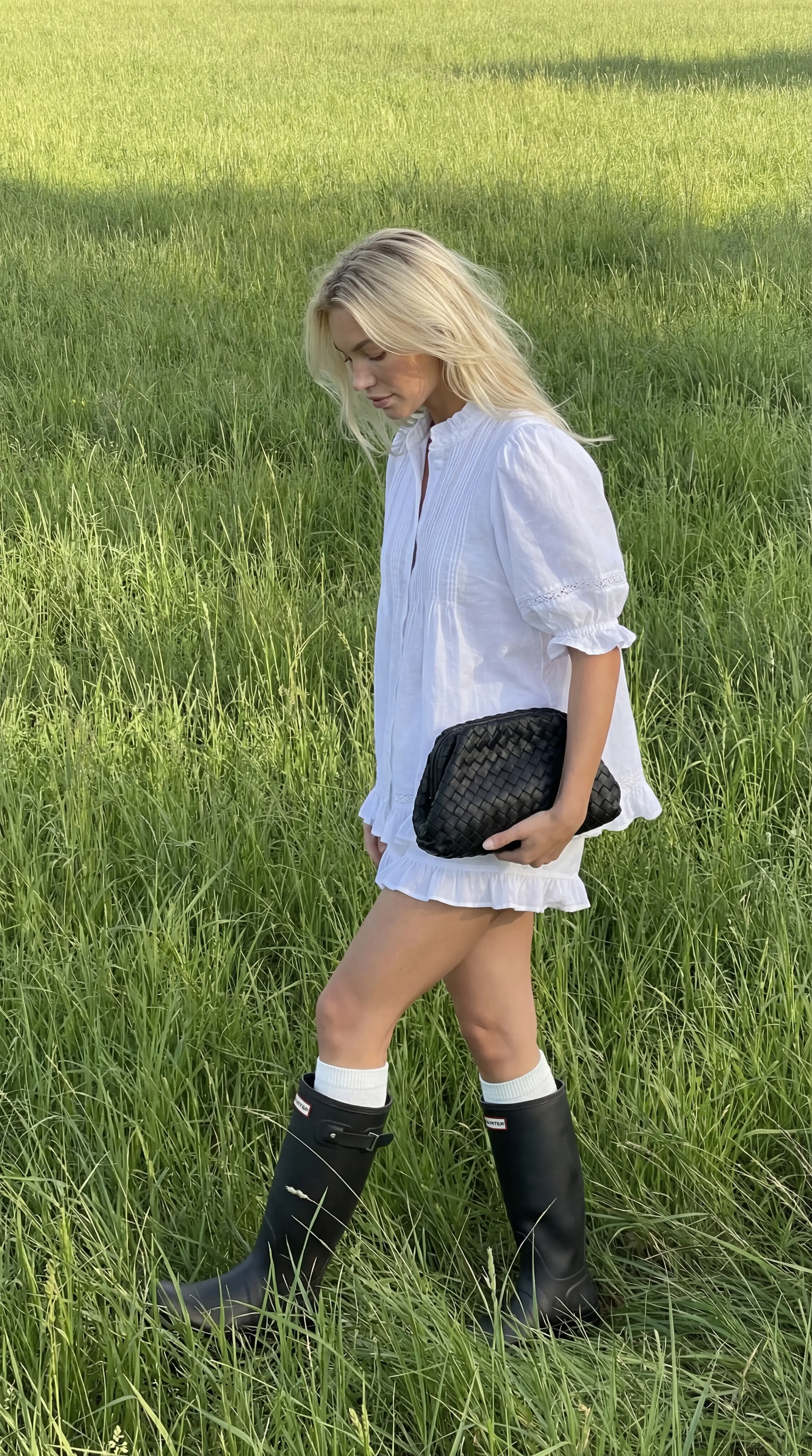 A woman with long blonde hair wearing a loose white dress, holding a black woven clutch, standing in a green grassy field.