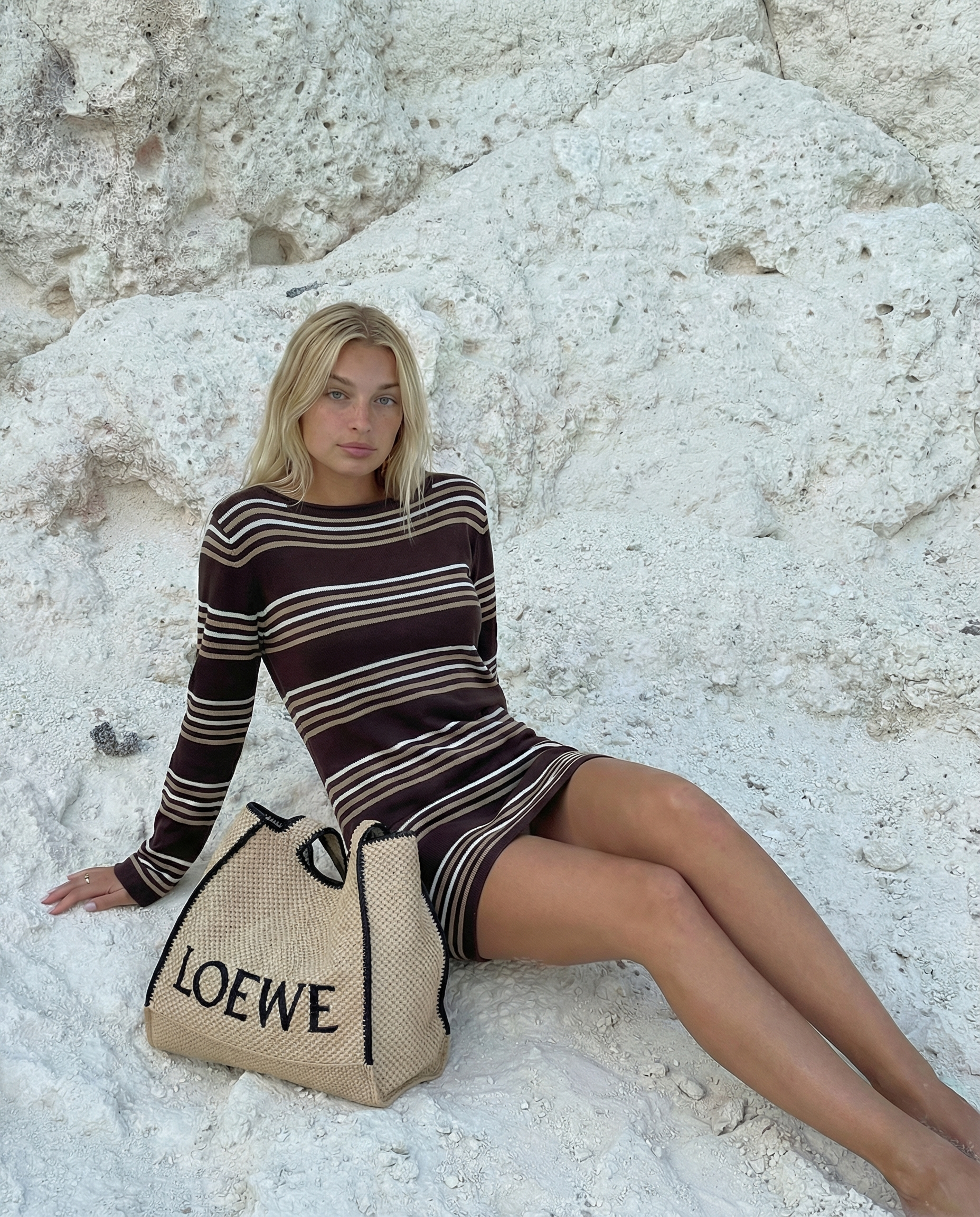 A woman in a striped dress sitting on white rocks near a cliffside with a Loewe beach bag beside her.