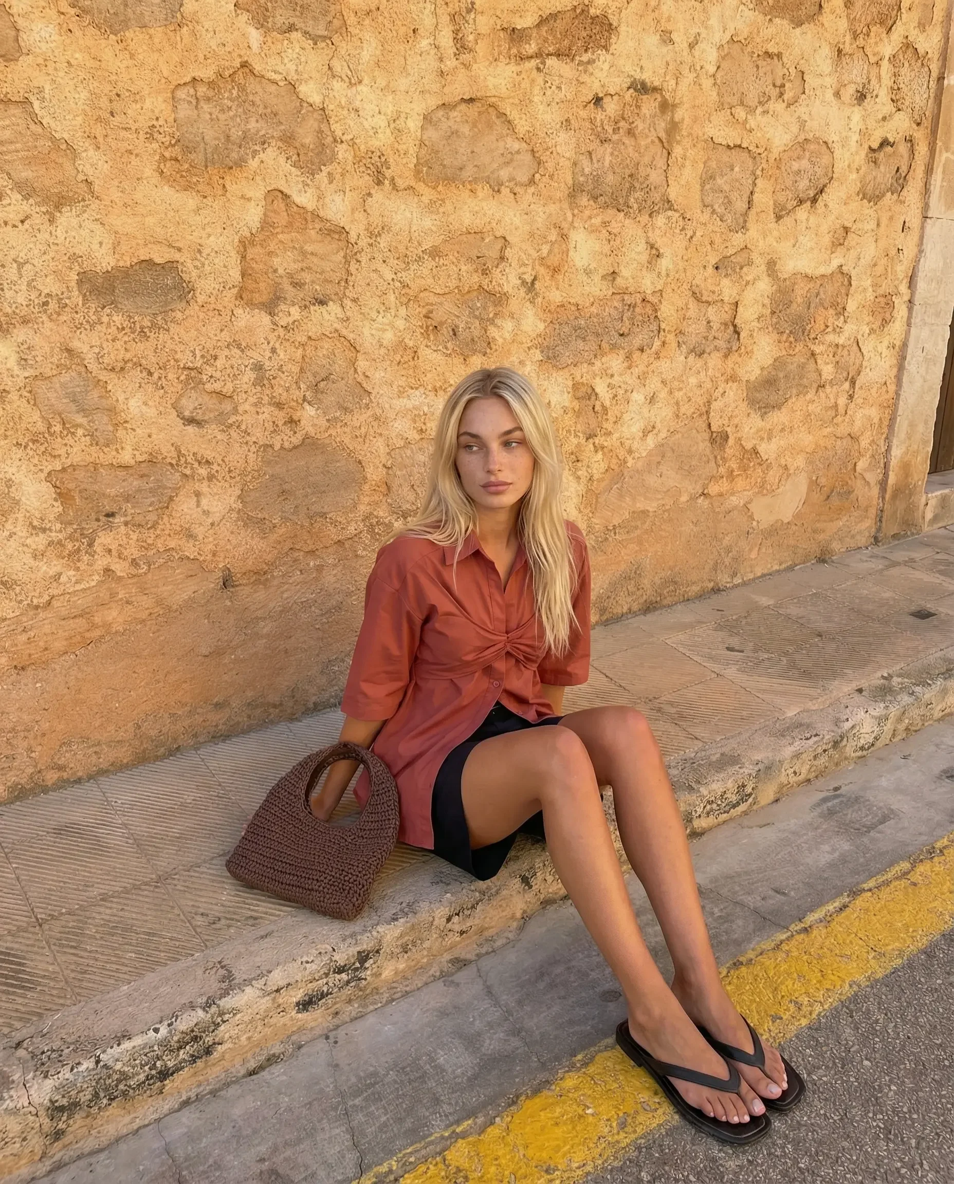 Young woman sitting on a sidewalk curb against a textured orange wall, wearing a rust-colored shirt, black shorts, and flip-flops, with a brown woven handbag beside her.