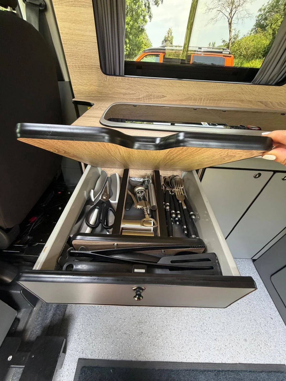 Open drawer in camper van kitchen containing various kitchen utensils and scissors, with window and trees visible outside.