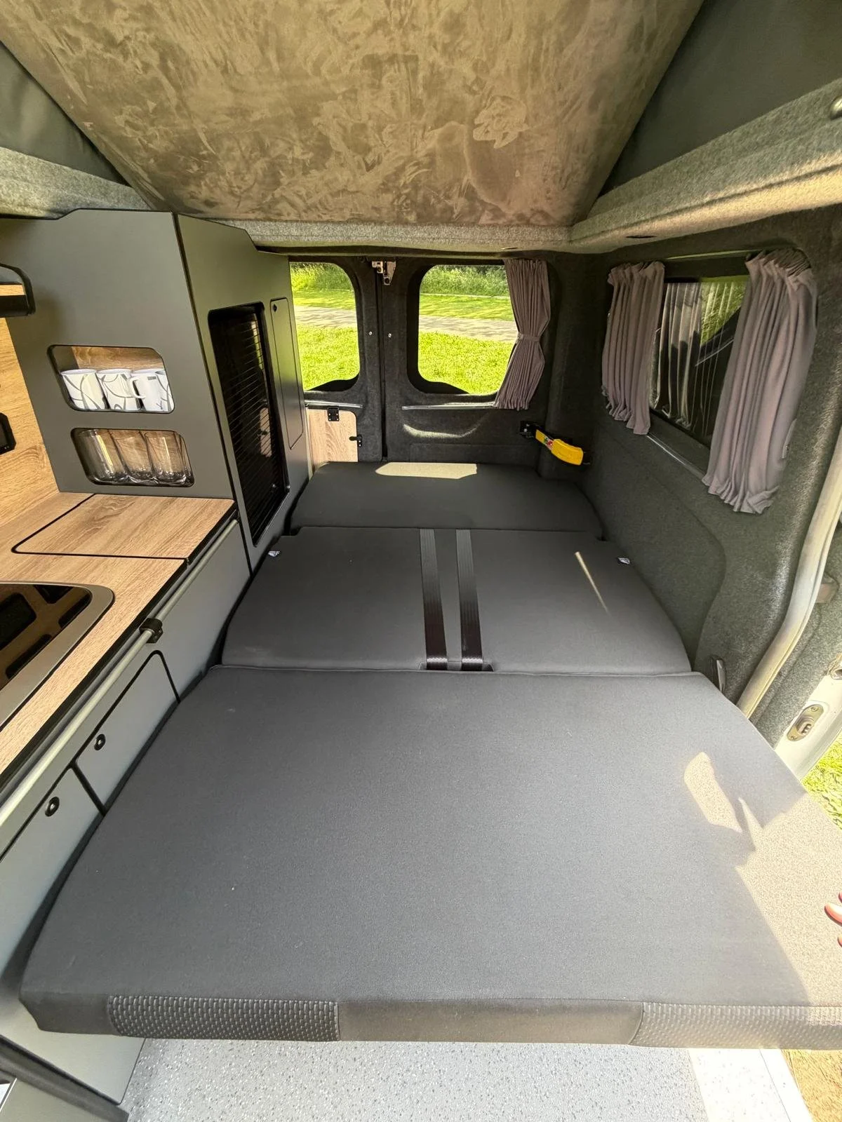 Interior of a camper van showing a sofa bed in the rear with windows and curtains, and a kitchenette with wooden countertops and cabinets on the left side.