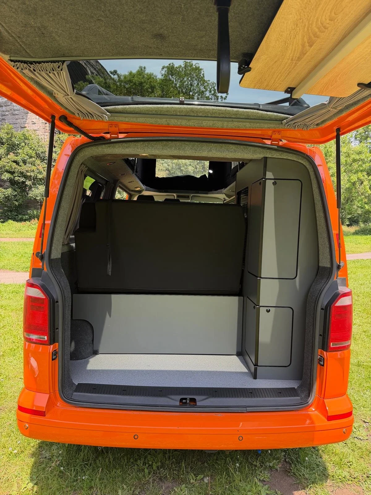 Open orange camper van showing storage compartments and interior, with a table attached to the ceiling, outdoors on grass and trees.