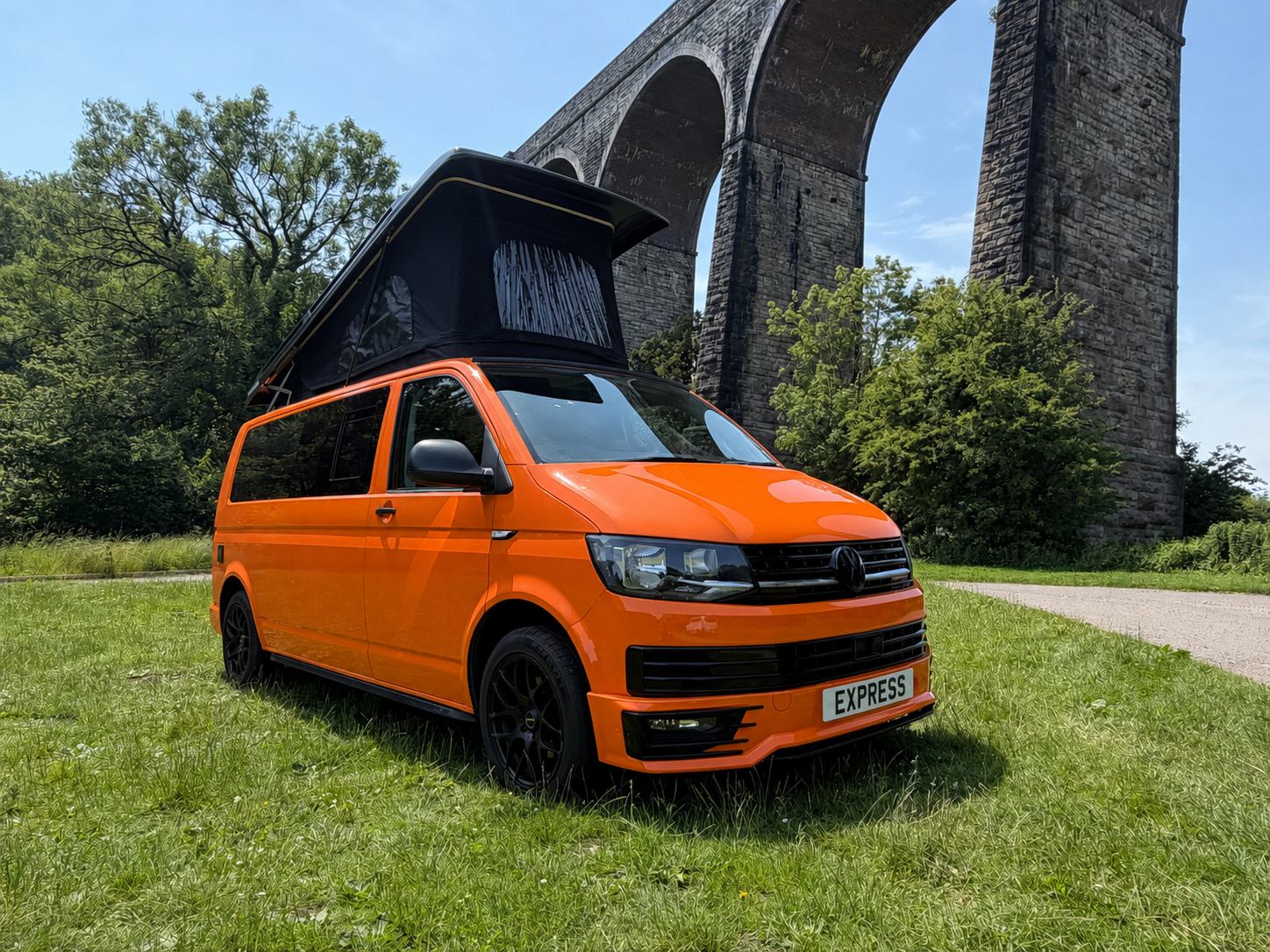 Orange camper van with a rooftop tent parked on a grassy area near a stone bridge under a blue sky.