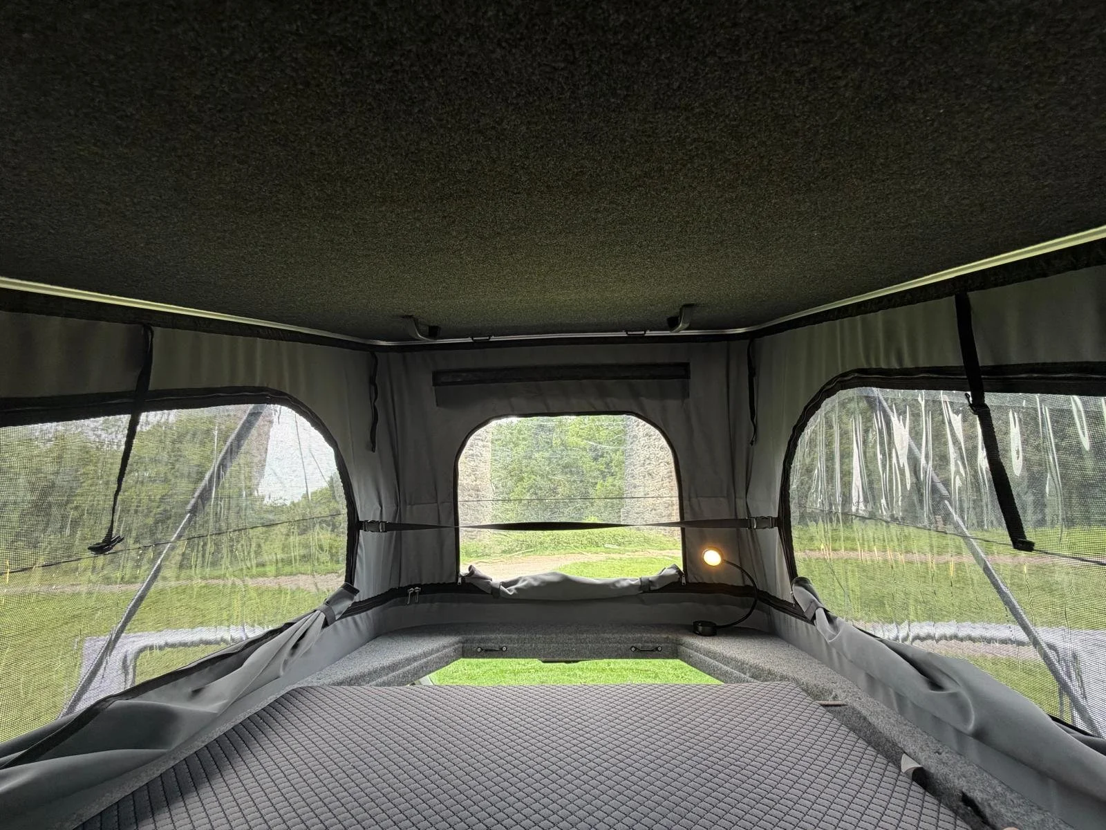 Interior view of a tent or camper with mesh windows, a screened door, and a fabric ceiling, overlooking a grassy outdoor area with trees in the background.