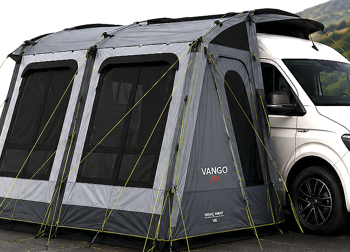 A white camper van with a large grey Vango Air camping tent attached to its side, set up outdoors on a paved area with hills in the background.