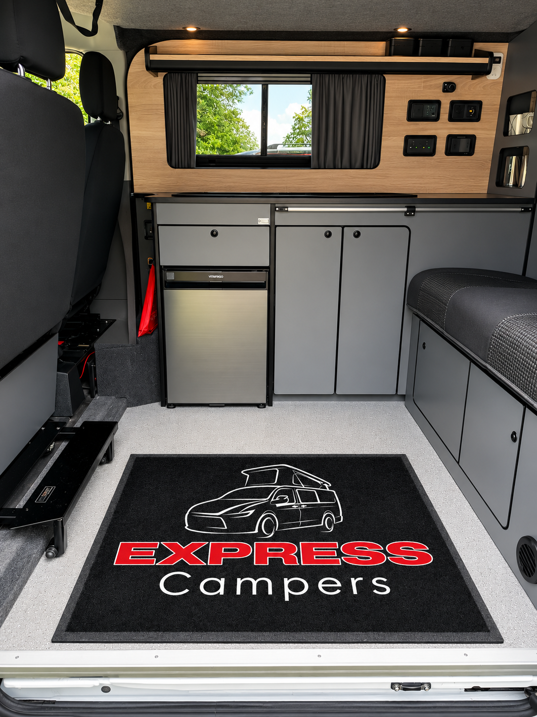 Interior view of a camper van with a black mat on the floor that says 'Express Campers' with a graphic of a camper van, a window with black curtains, and various storage compartments and electrical outlets on wooden paneling.