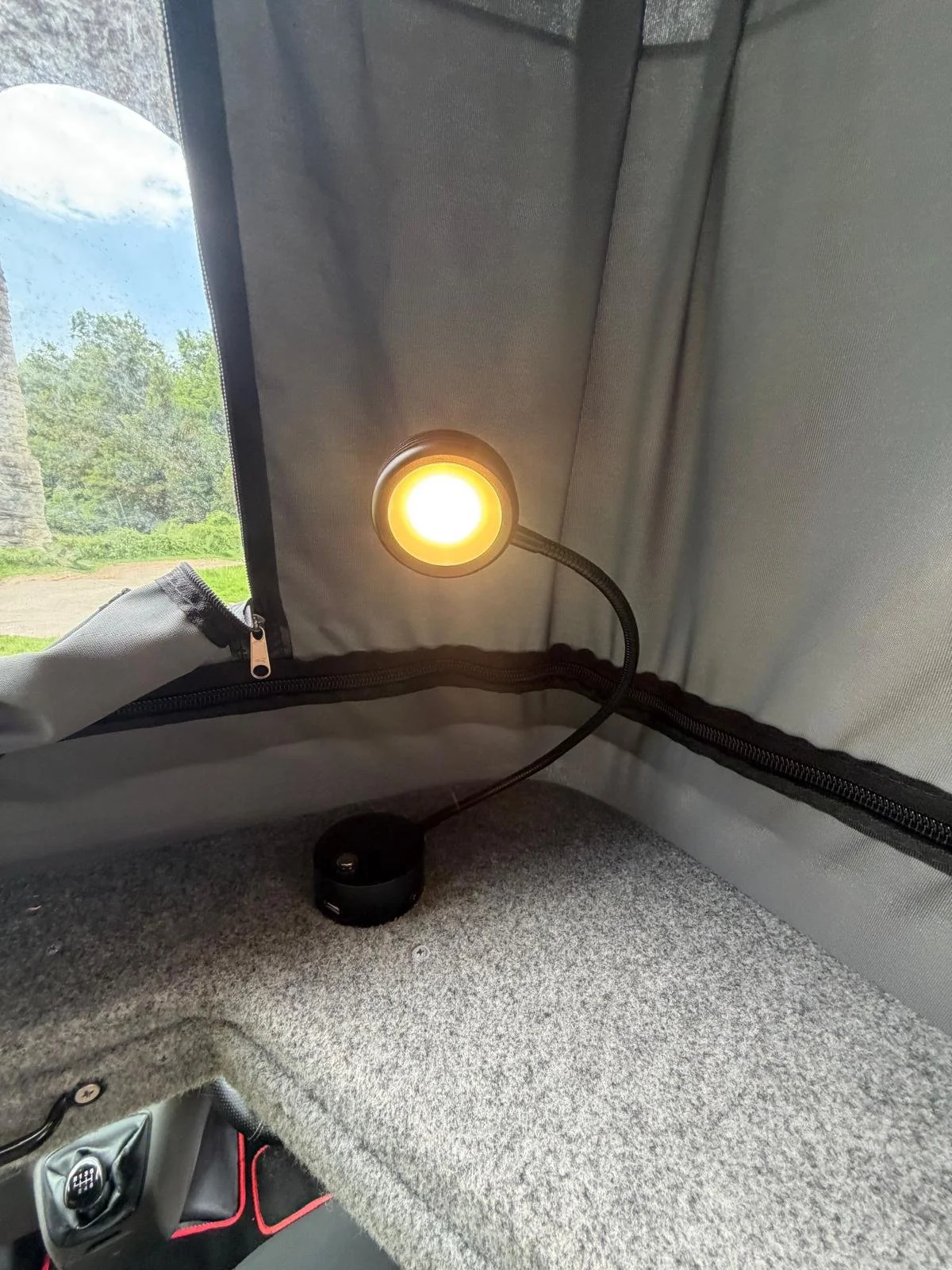 Inside a vehicle, a small round portable LED light with a flexible gooseneck is turned on, emitting warm light near the back passenger side, with a zippered compartment and a view of outdoors through a window.
