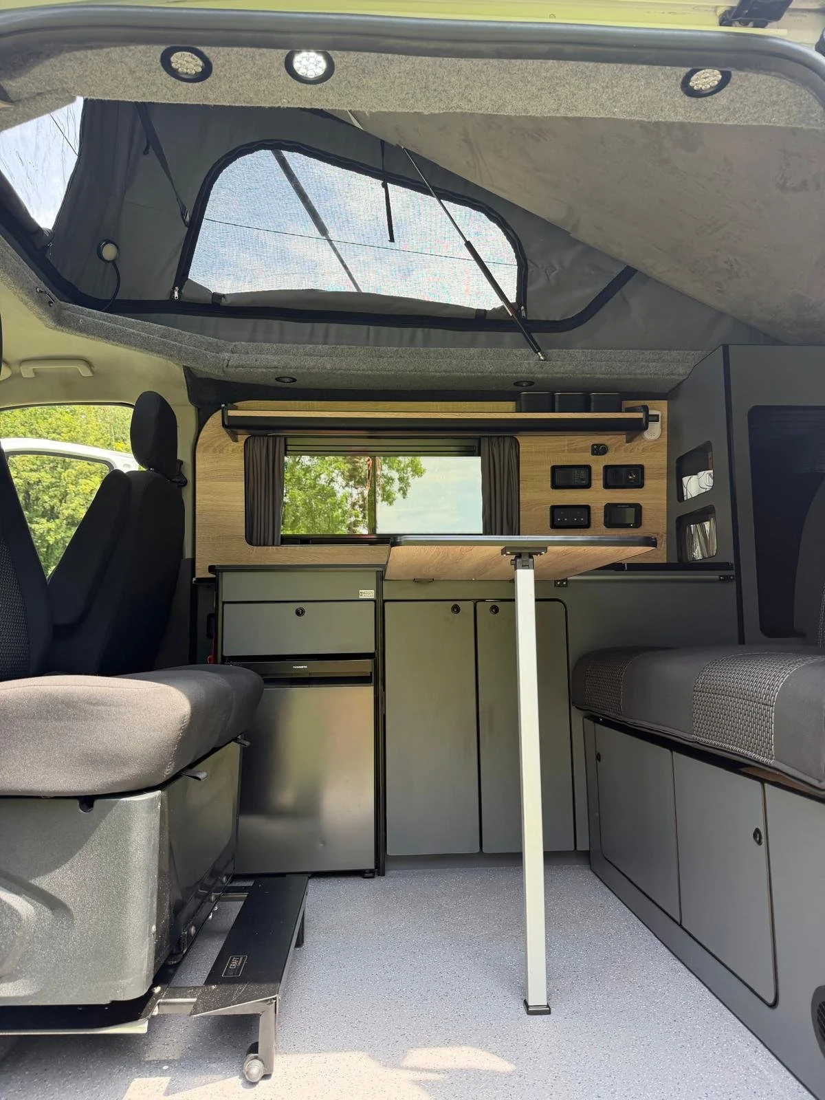Interior of a camper van with seating area, small fridge, wooden wall with electrical outlets, window with curtains, and a ceiling window.